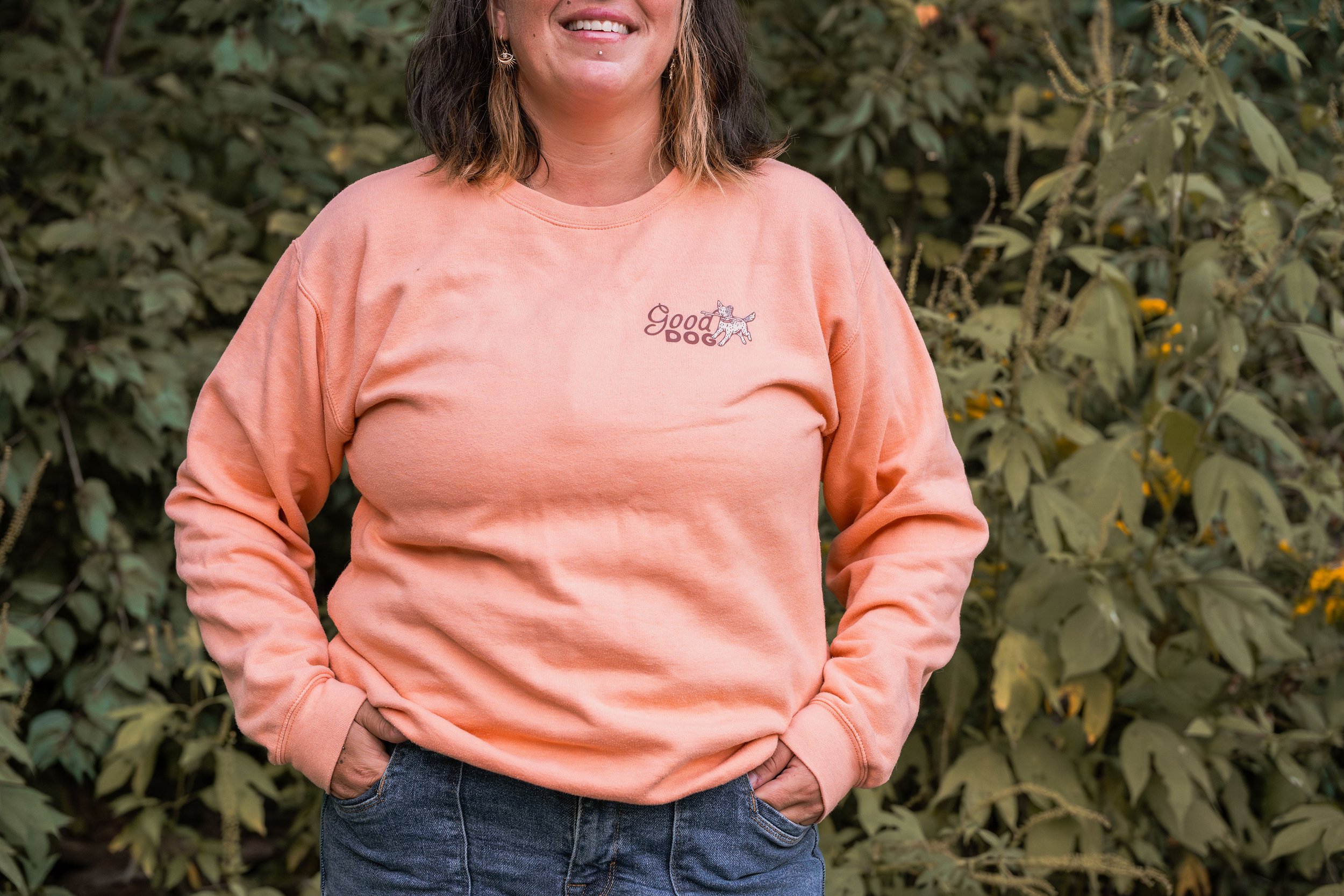 A person with shoulder-length brown hair, wearing a peach-colored sweatshirt with a small graphic of a dog and the words 'Good Dog' on the chest, standing outdoors in front of green foliage.