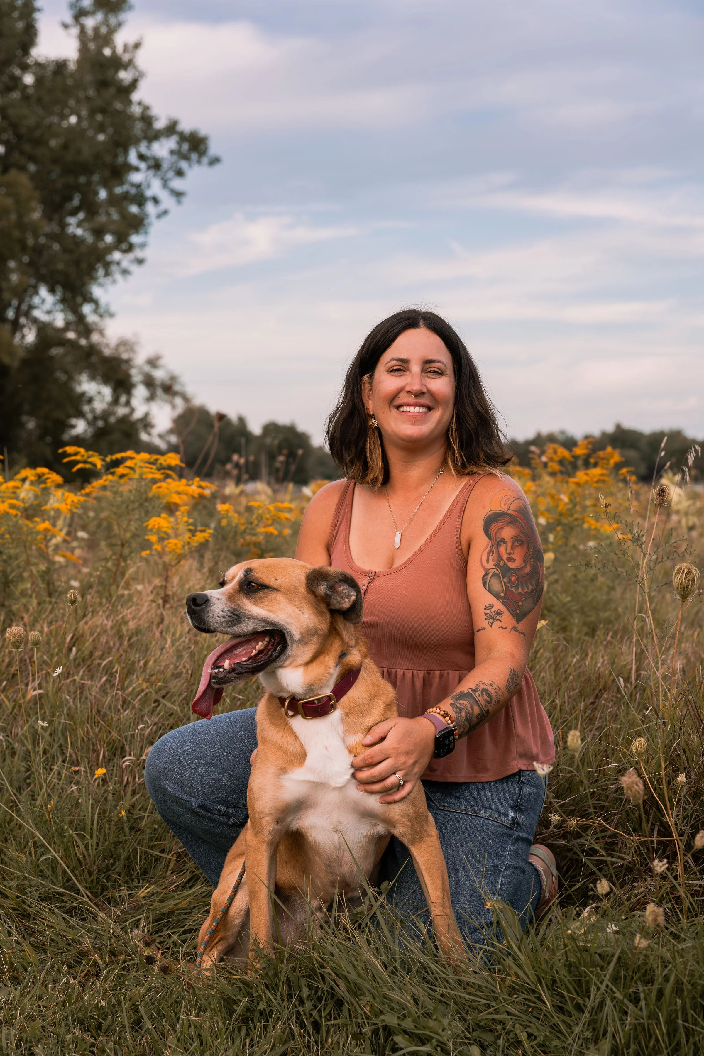 A woman with tattoos smiling while sitting outdoors with a brown and white dog in a field of yellow flowers, under a partly cloudy sky.