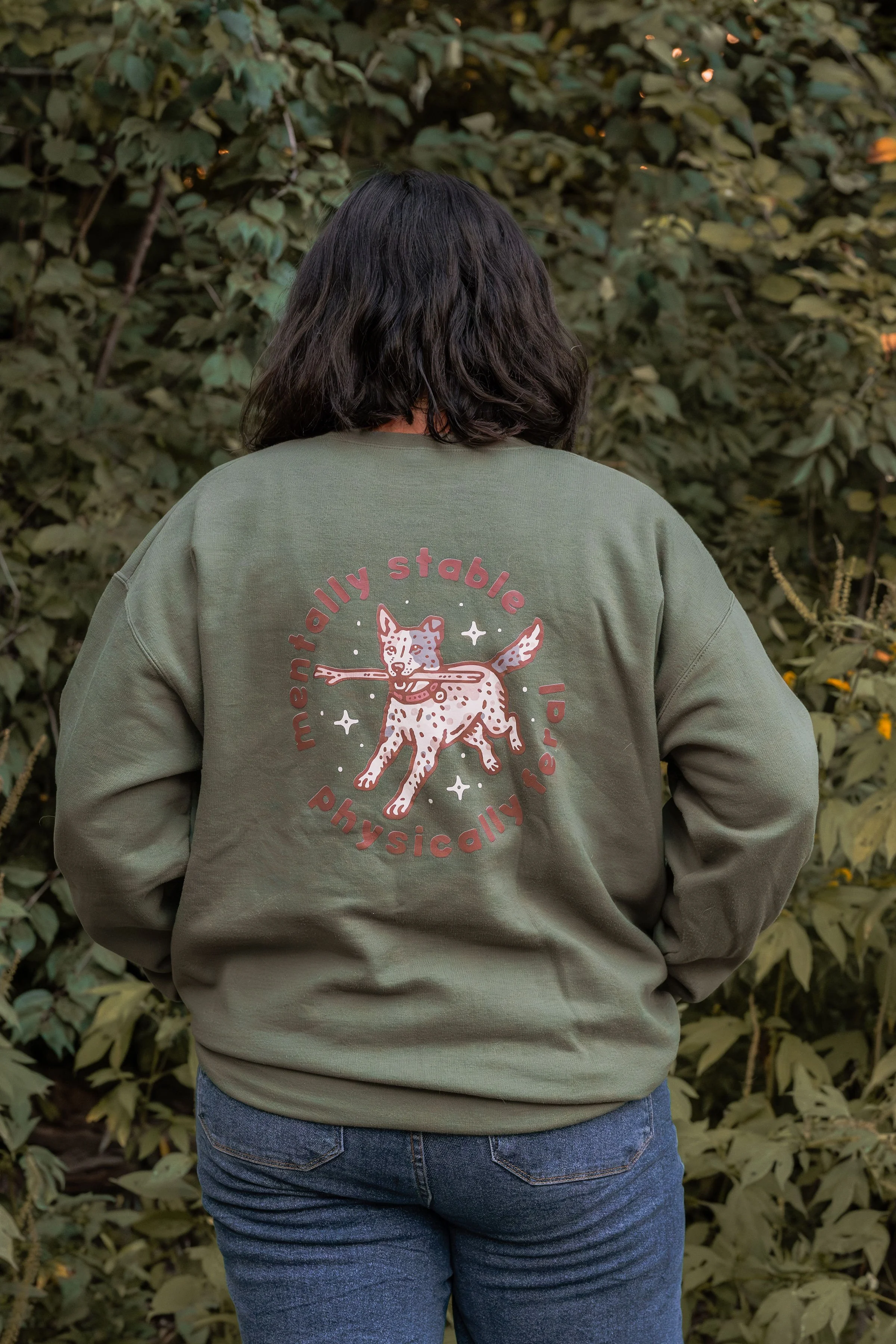 Back view of a person with shoulder-length black hair wearing a green sweatshirt with a graphic of a dog and the words 'mentally stable physically related' on the back. The person is standing in front of a leafy shrub.
