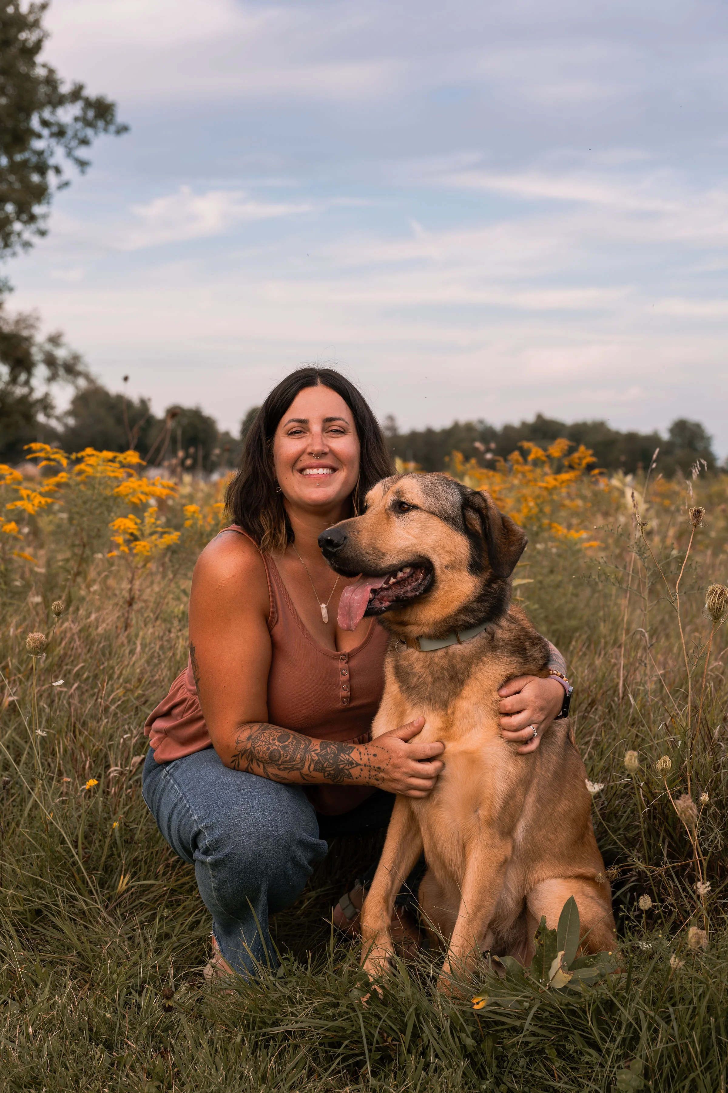 A woman with dark hair, tattoos on her arms, and a nose piercing smiling and sitting in a field of wildflowers with a large tan and black dog that has its tongue out.