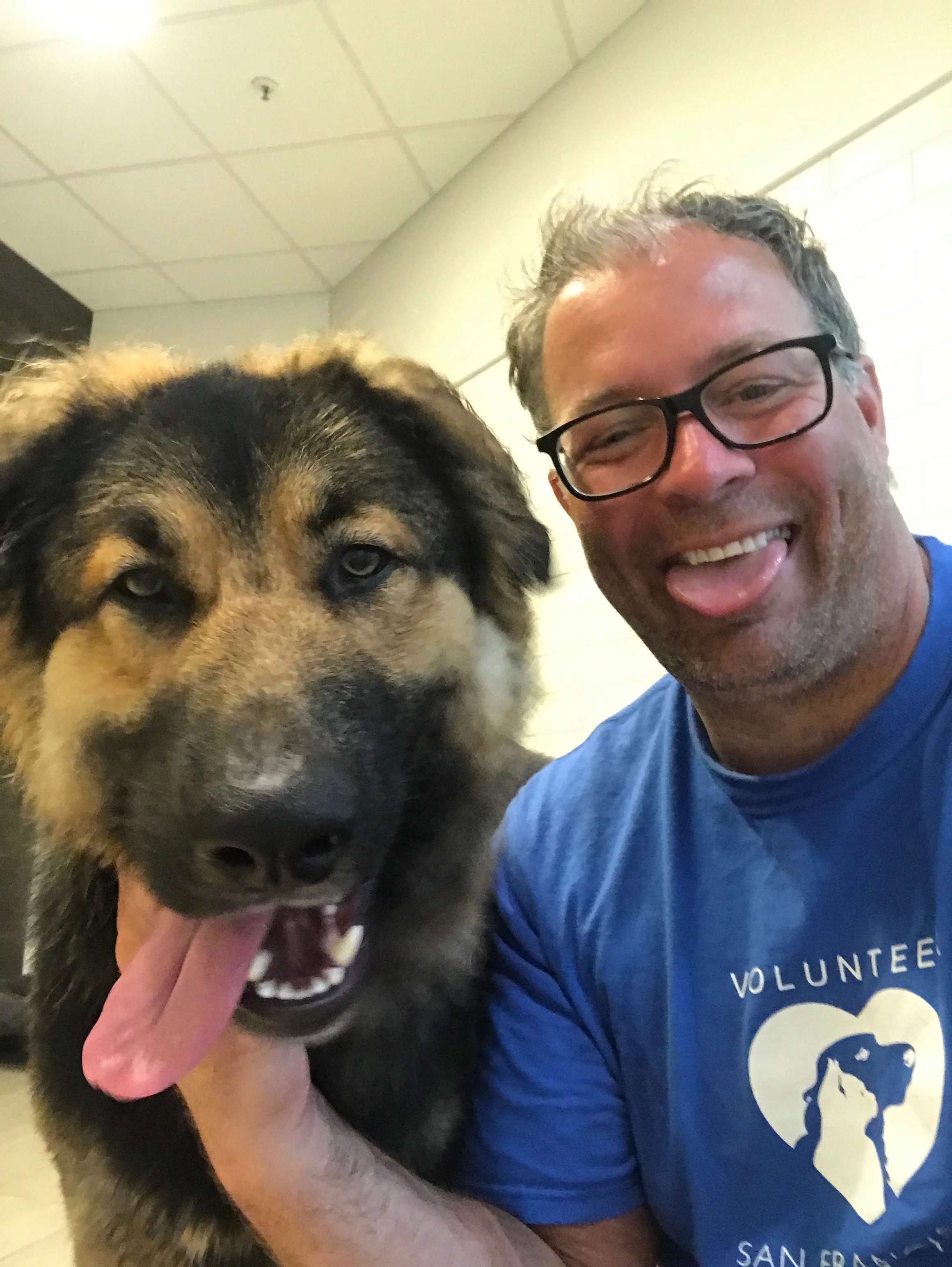 A man with glasses smiling and sticking out his tongue, posing with a large, fluffy dog with black fur and tan markings, inside a building with a ceiling and lights visible.