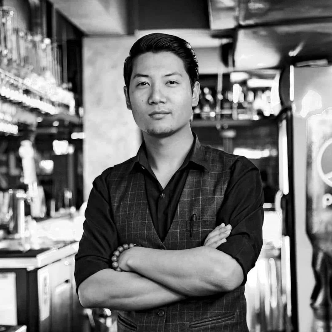 Black and white photo of a young man with crossed arms, wearing a vest and shirt, standing in a bar or restaurant.