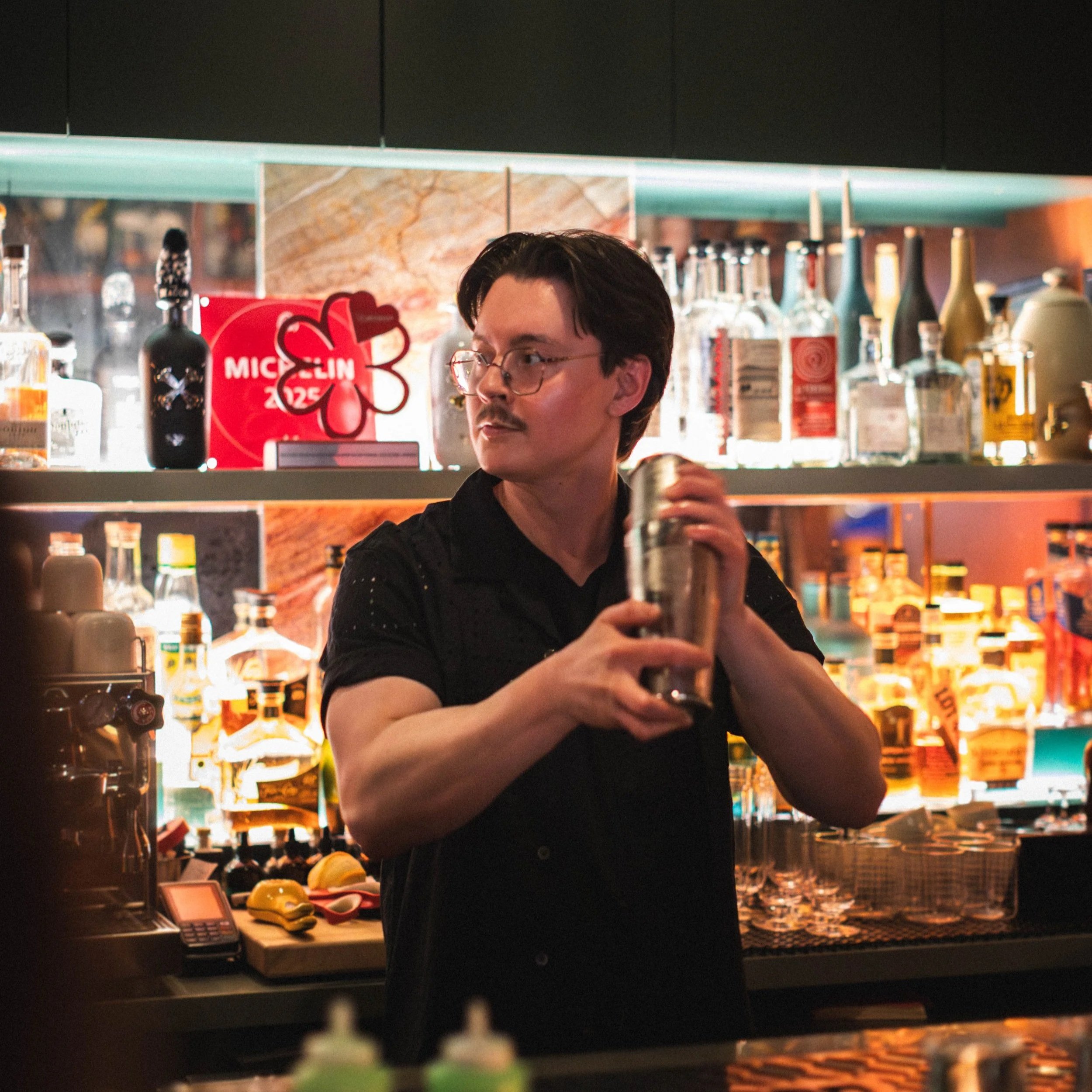 A man with dark hair and glasses at a bar or restaurant, with bottles, a cash register, and a red sign with a flower design and the word 'Michelin' in the background.