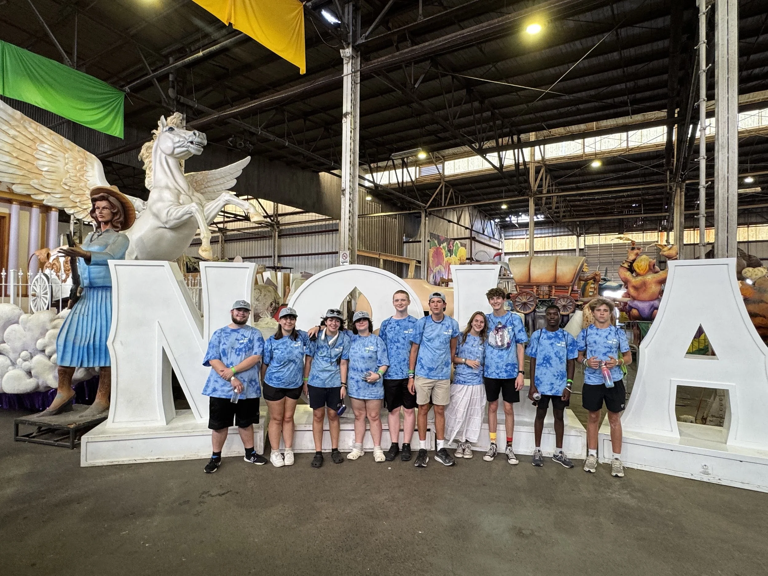 Group of nine young people standing in front of a large ornate sculpture of a winged horse and a woman in a warehouse-like setting with high ceilings.