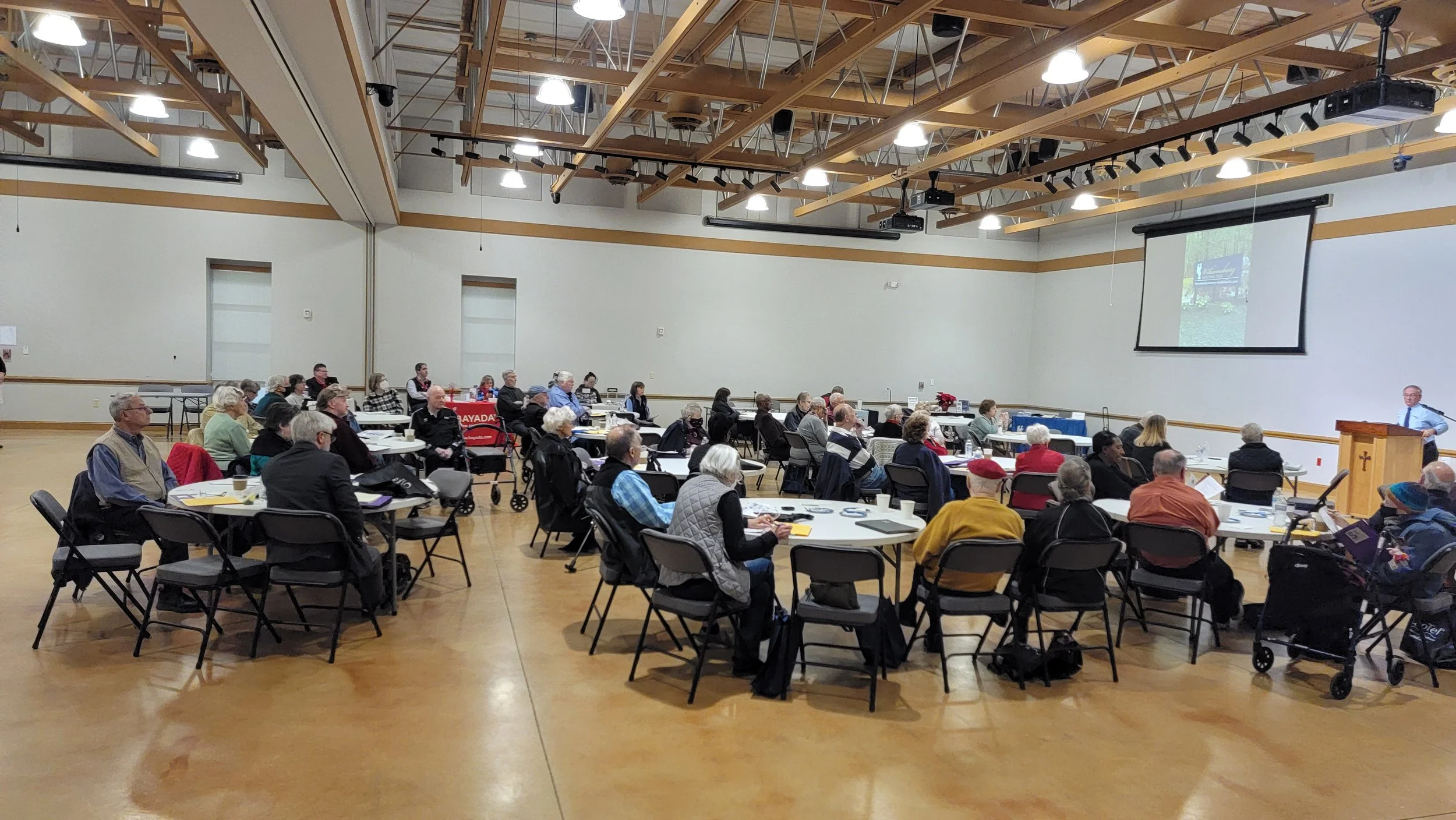 A large group of people attending a presentation or conference in a spacious room with wooden ceiling beams, a projector screen, and a speaker at a podium.