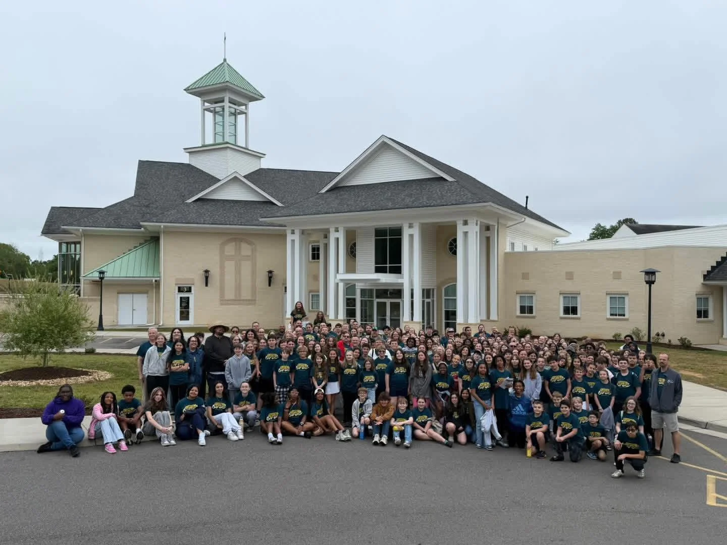 Large group of people, including children and adults, gathered in front of a multi-story building with white columns and a clock tower, during daytime, on a cloudy day.