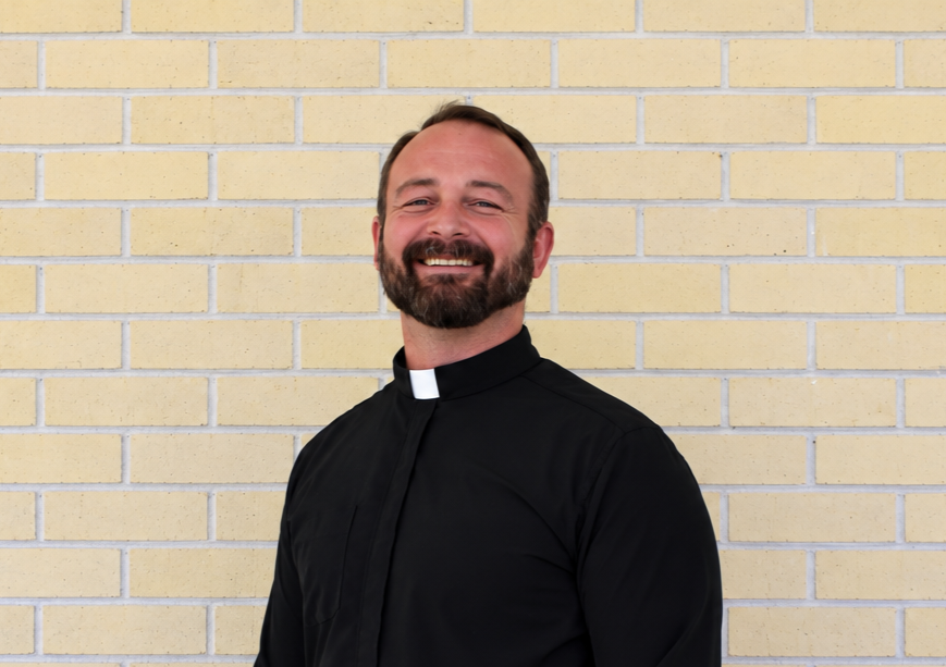 A smiling man with a beard and short hair wearing a priest's collar, standing against a beige brick wall.
