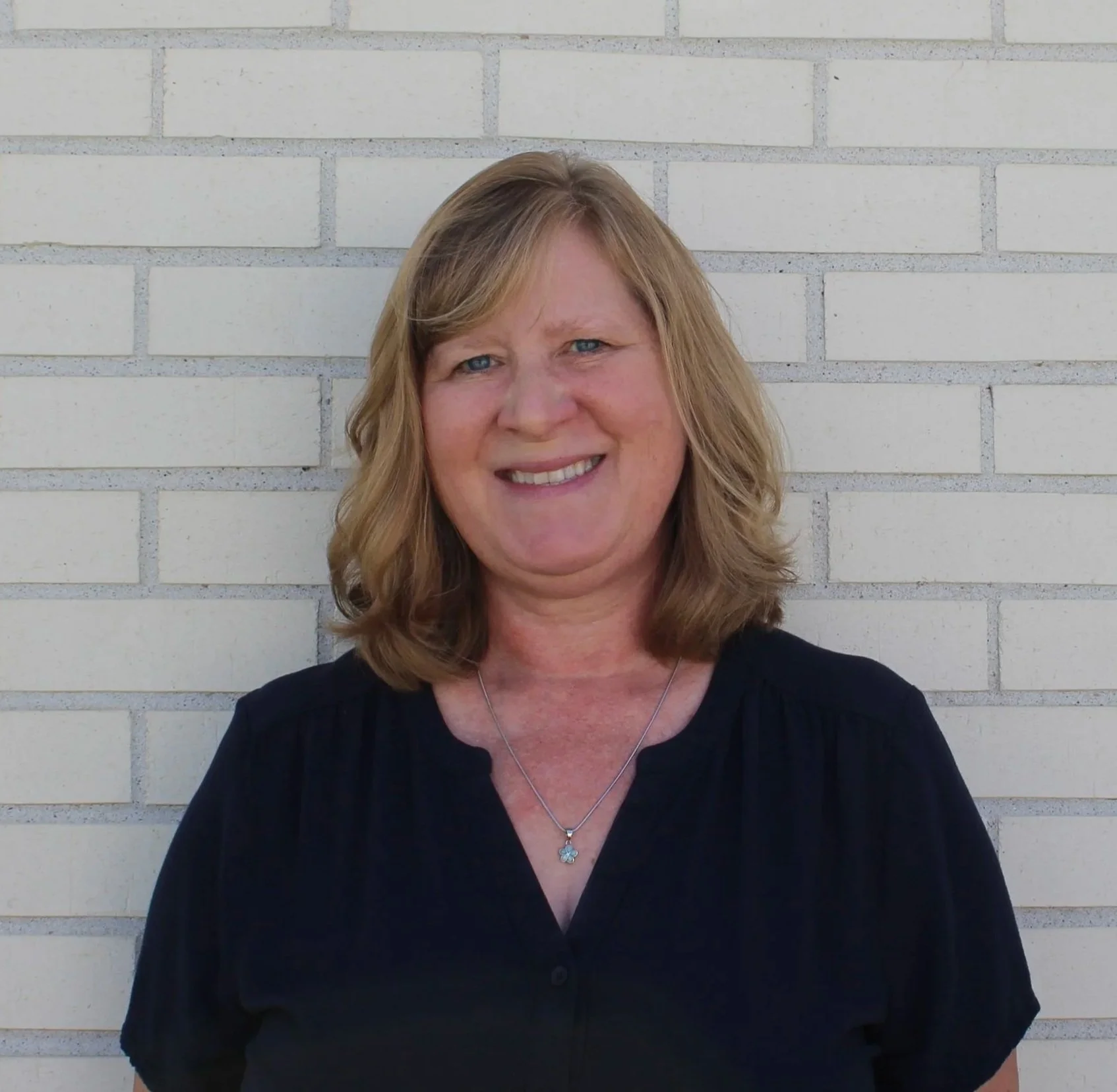 A woman with shoulder-length blonde hair and blue eyes smiling, wearing a black top and a silver necklace with a four-leaf clover pendant, standing in front of a light-colored brick wall.