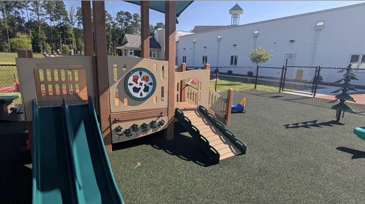 Empty playground with a slide, bridge, and climbing structure, enclosed by a fence, with a building and trees in the background.