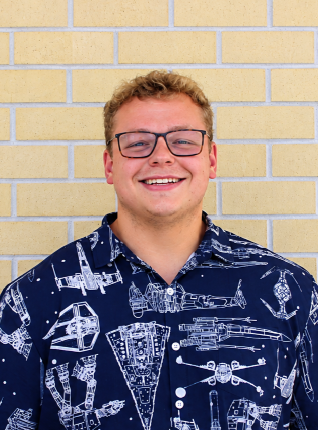 A young man with curly blond hair, wearing glasses and a navy blue shirt with white spaceship prints, smiling in front of a yellow brick wall.