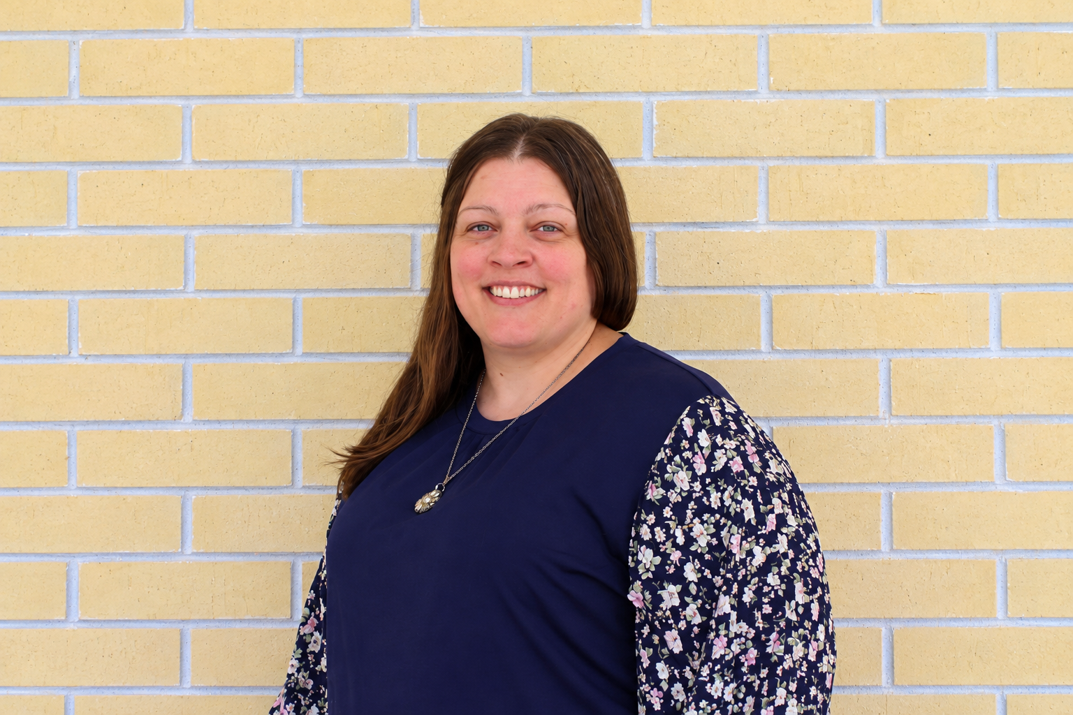 A woman with long brown hair and a big smile standing against a yellow brick wall, wearing a navy blue top with floral-patterned sleeves and a silver necklace.