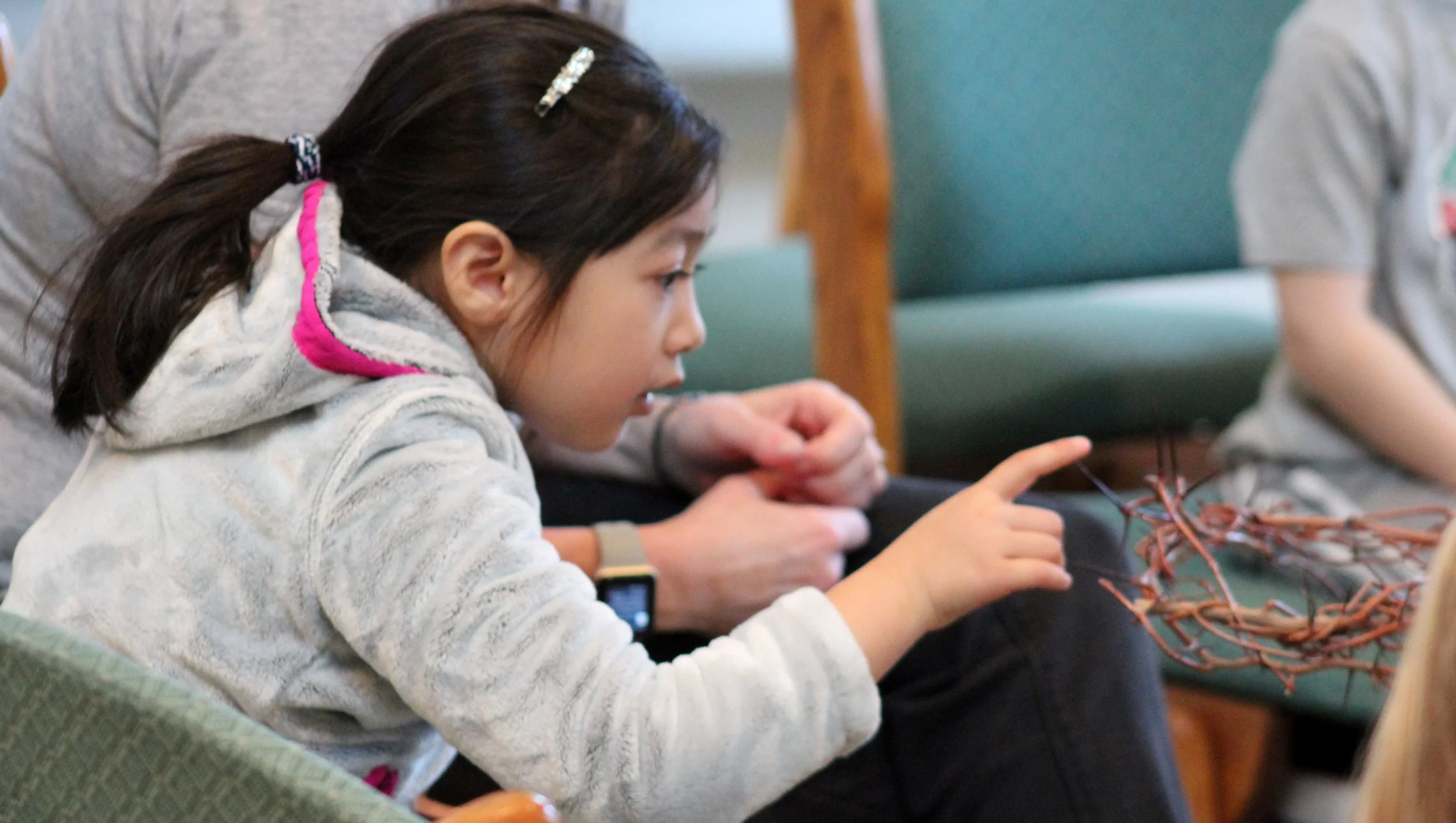 Young girl with long dark hair, wearing a hair clip and a white hoodie, sitting on a green couch, using a smartphone, with a person beside her holding a bundle of twigs.