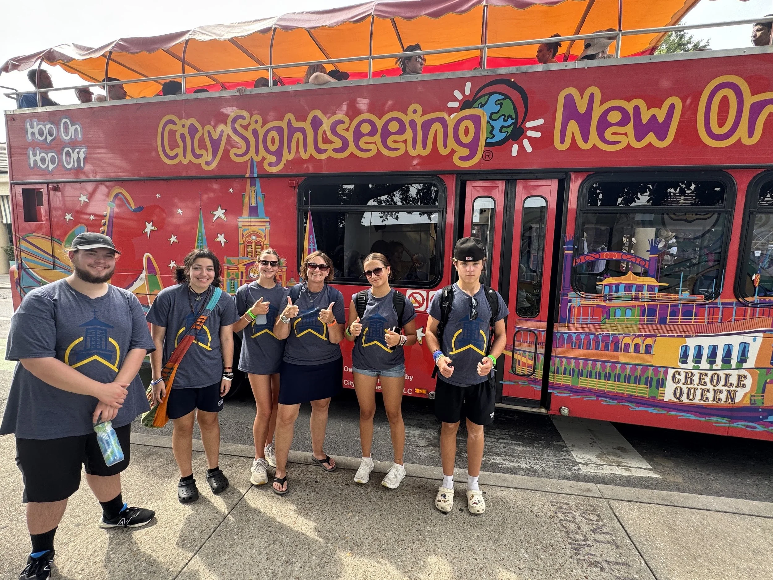 A group of six people standing in front of a colorful city sightseeing double-decker bus. The people are smiling, giving thumbs up, and wearing matching dark gray T-shirts. The bus is decorated with vibrant illustrations of a roller coaster and a cas