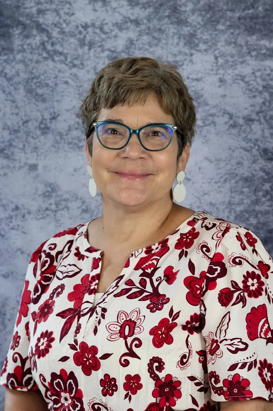 A woman with short brown hair, glasses, and white earrings smiling in front of a gray mottled background, wearing a white blouse with red and black floral pattern.