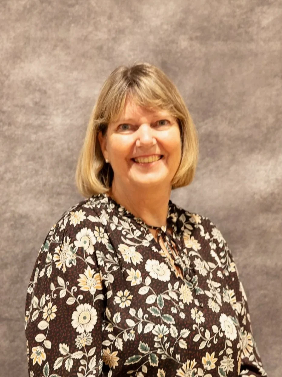 A smiling woman with shoulder-length blond hair, wearing a floral patterned blouse, standing against a neutral background.