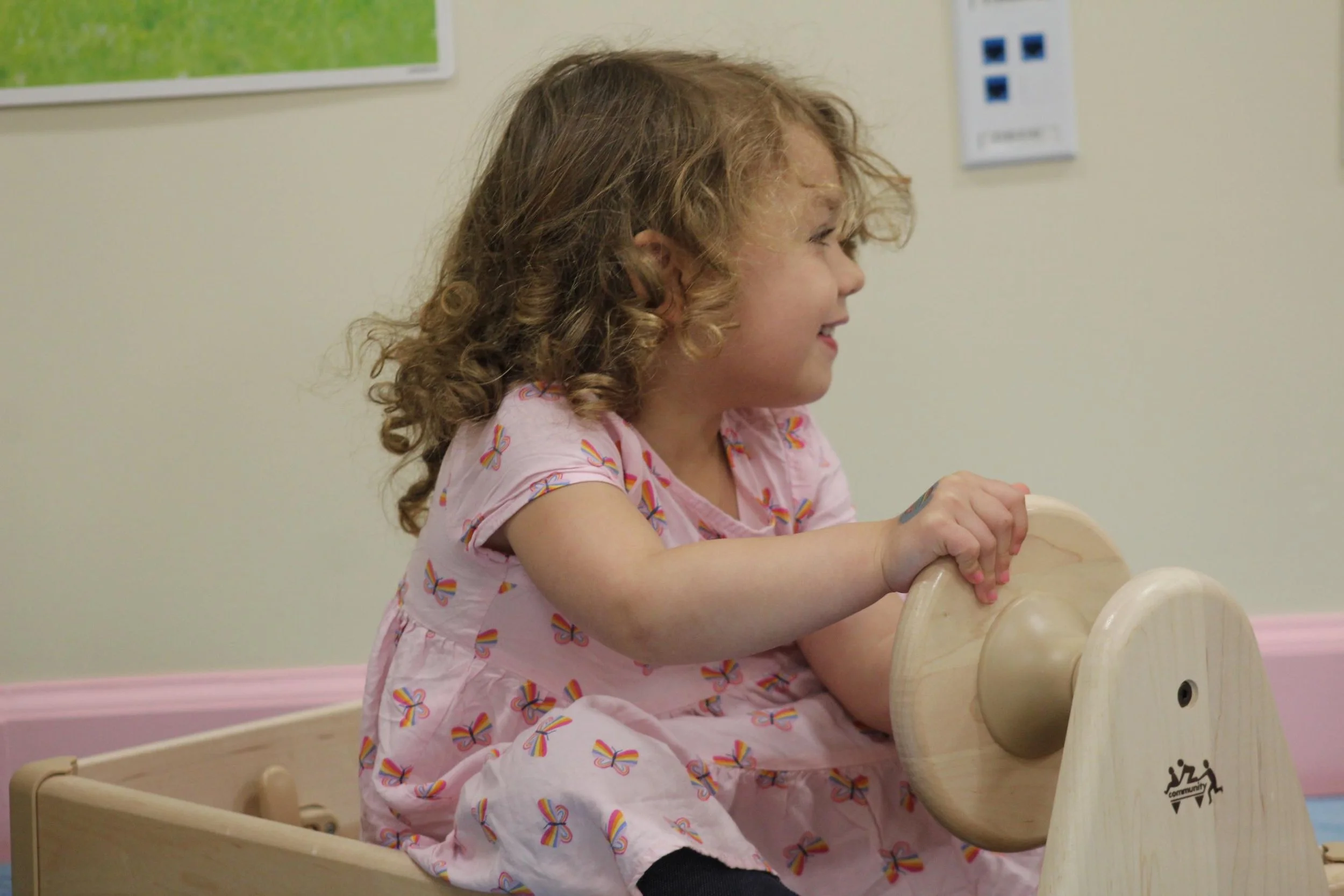 A young girl with curly red hair wearing a pink dress with rainbow print, sitting on a wooden rocking horse, smiling.