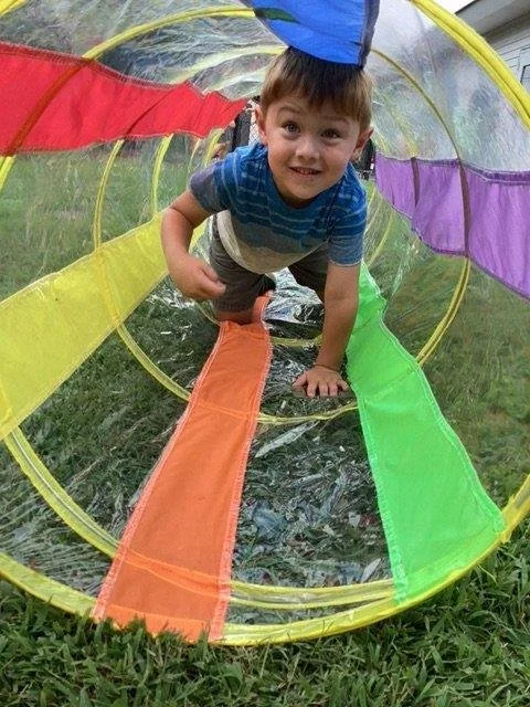 A young boy crawling through a colorful, collapsible play tunnel outdoors on grass, looking at the camera and smiling.