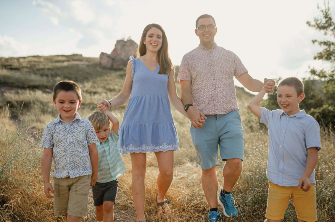A family of five walking outdoors through a grassy field during daytime, holding hands and smiling.