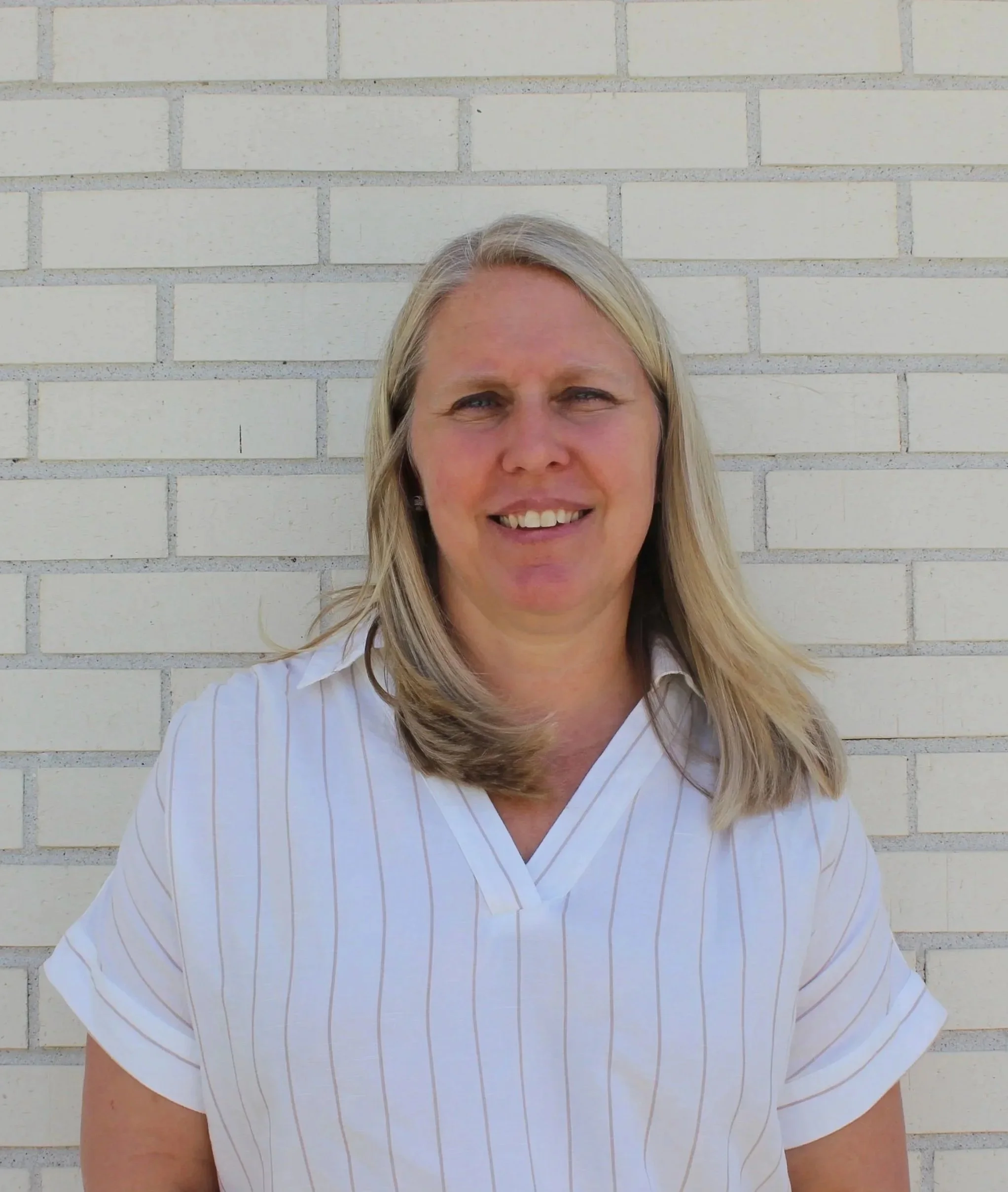 Close-up of a woman with long blond hair, smiling, standing against a white brick wall. She is wearing a white, short-sleeved, collared shirt with thin pinkish stripes.