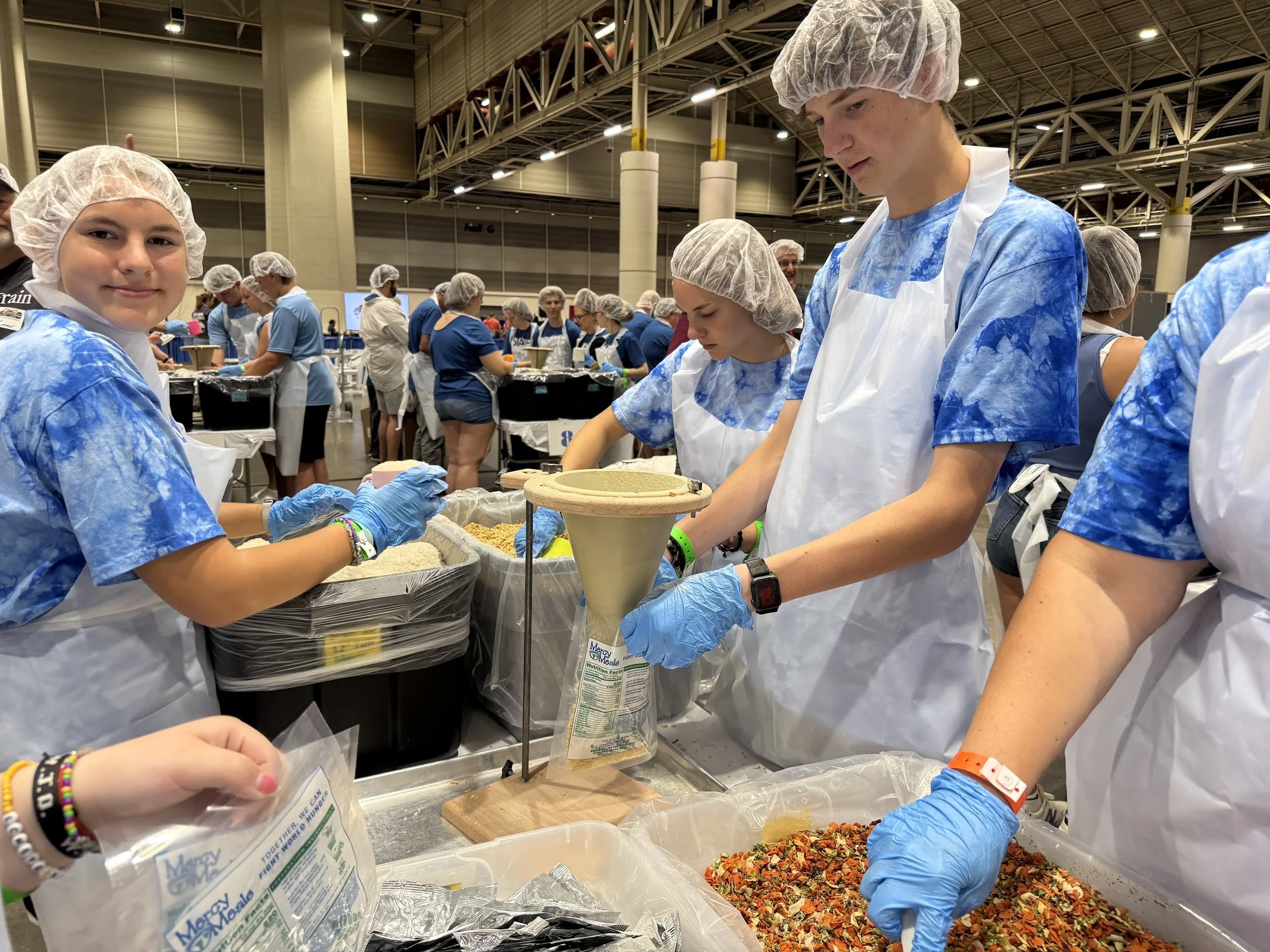 Teenagers wearing hairnets, face masks, and aprons working at a food service station in a large indoor space, preparing food with various ingredients.