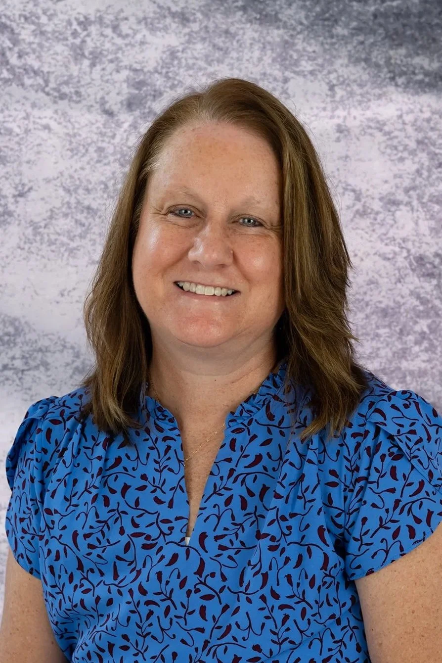 A woman with shoulder-length brown hair, blue eyes, and fair skin, smiling, wearing a blue blouse with a dark leaf pattern, standing against a textured gray background.