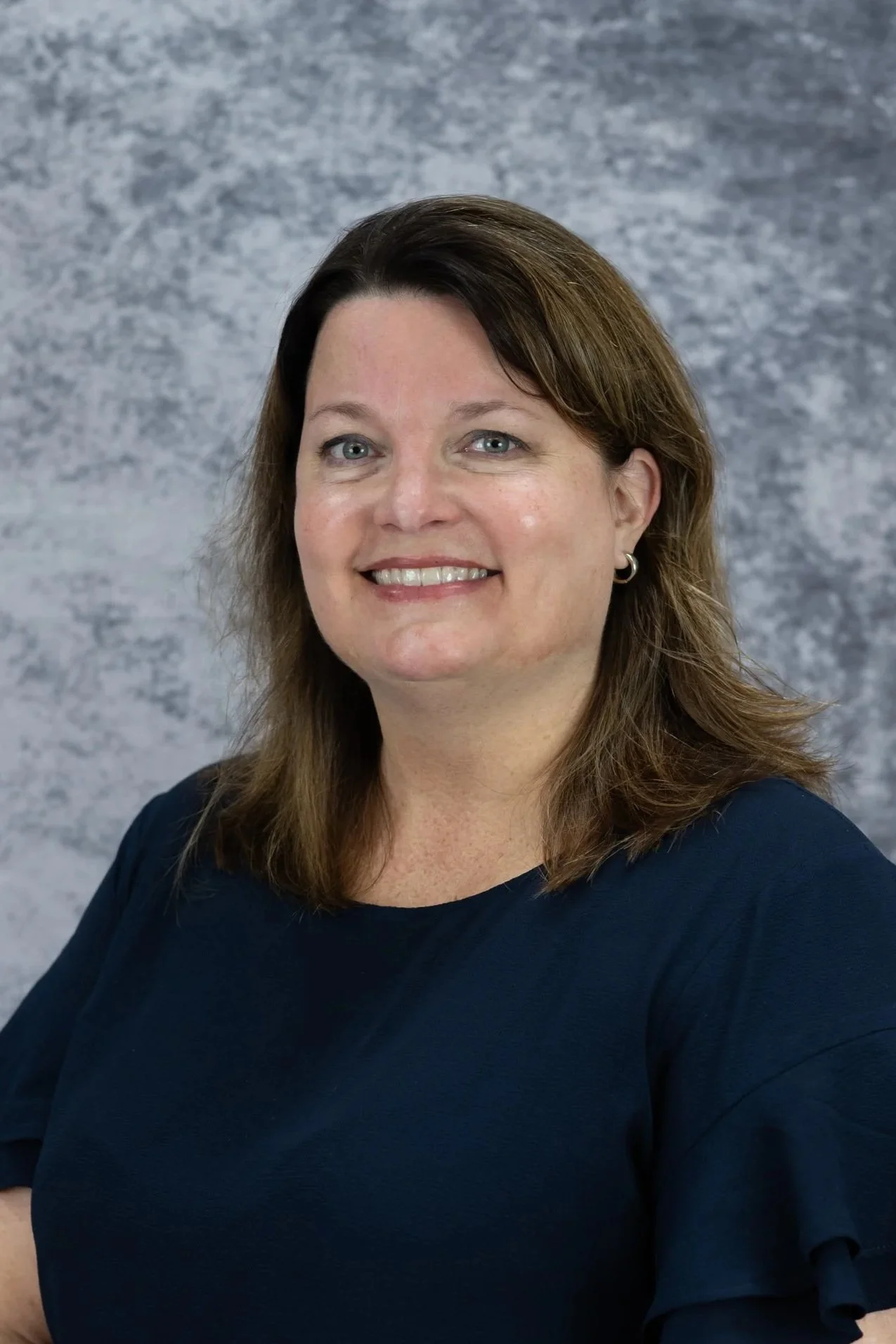 A woman with shoulder-length brown hair, blue eyes, and earrings, smiling and wearing a dark blue top, standing against a gray mottled background.