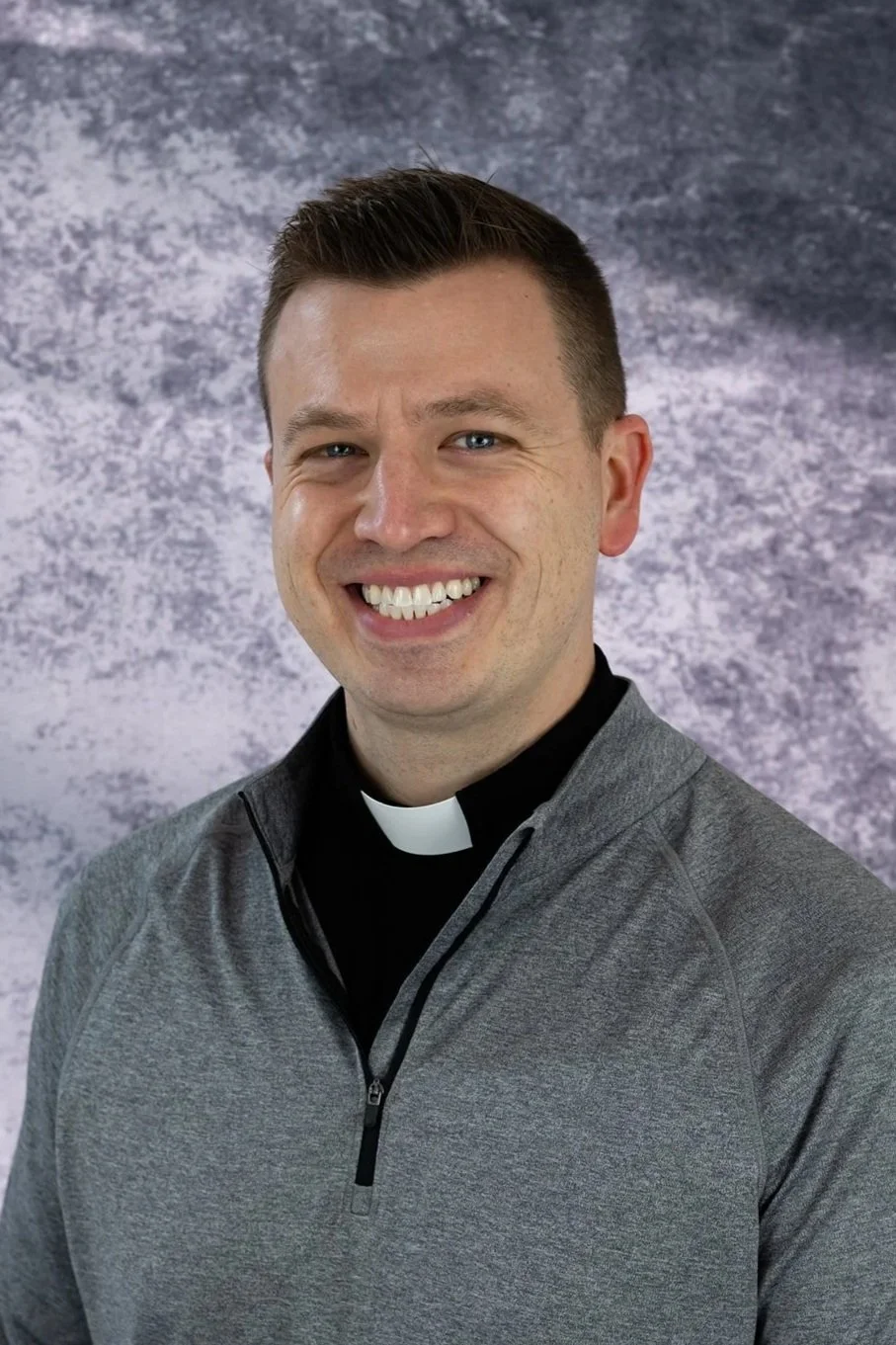 Portrait of a smiling man dressed as a priest, wearing a clerical collar and a gray athletic jacket, against a textured purple and gray background.