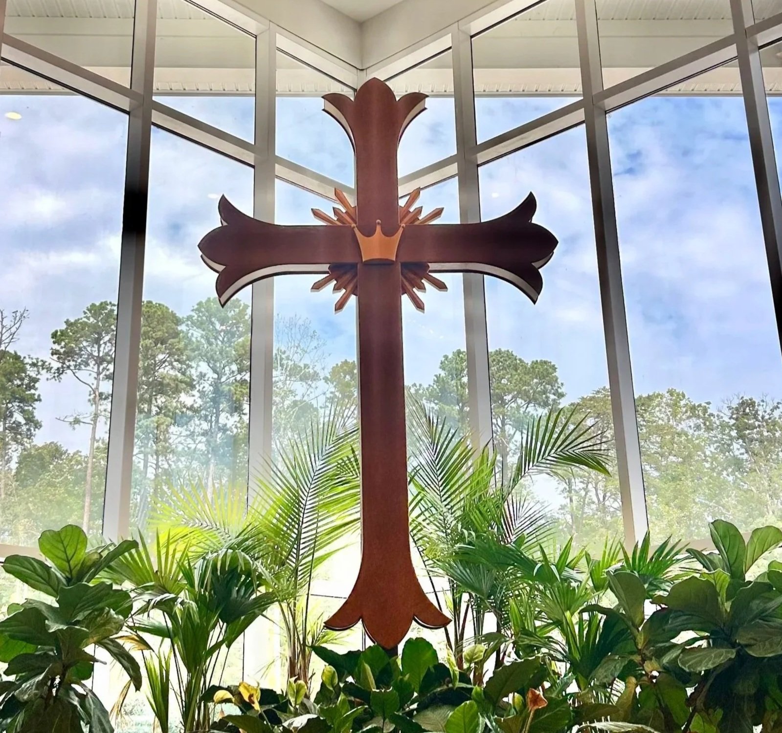 A wooden cross with a crown and rays design behind it, surrounded by lush green plants, with large windows showcasing trees and a blue sky in the background.