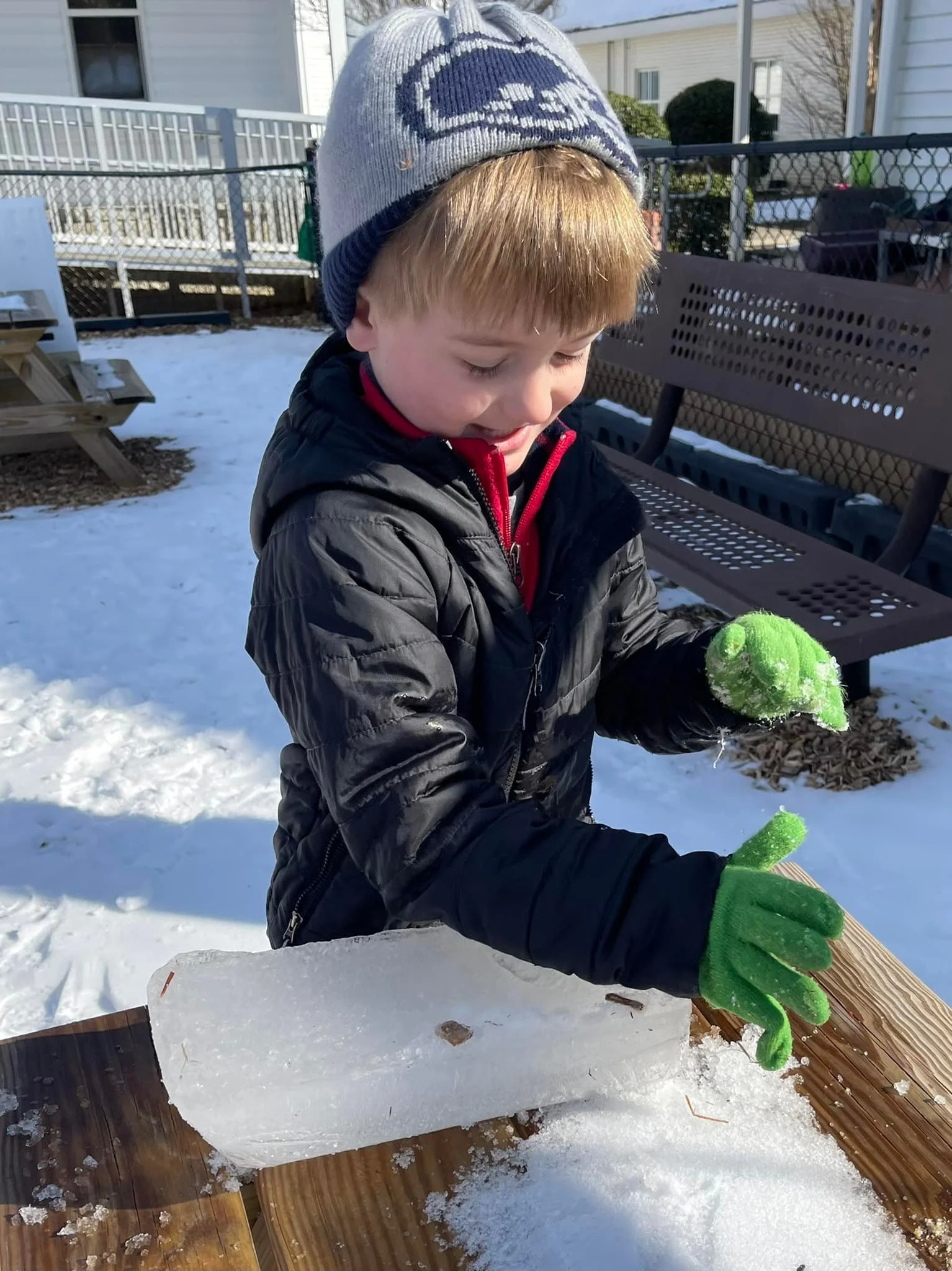 A young boy wearing a grey and blue beanie, a black jacket, and green gloves, is playing with a snow-covered wooden object outdoors in a snowy backyard.