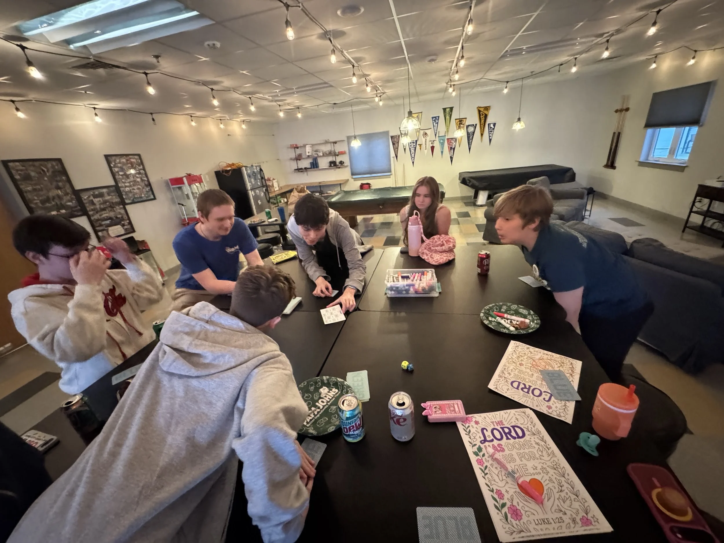 People gathered around a table playing a game, with drinks and game materials visible, in a decorated room with string lights and banners.