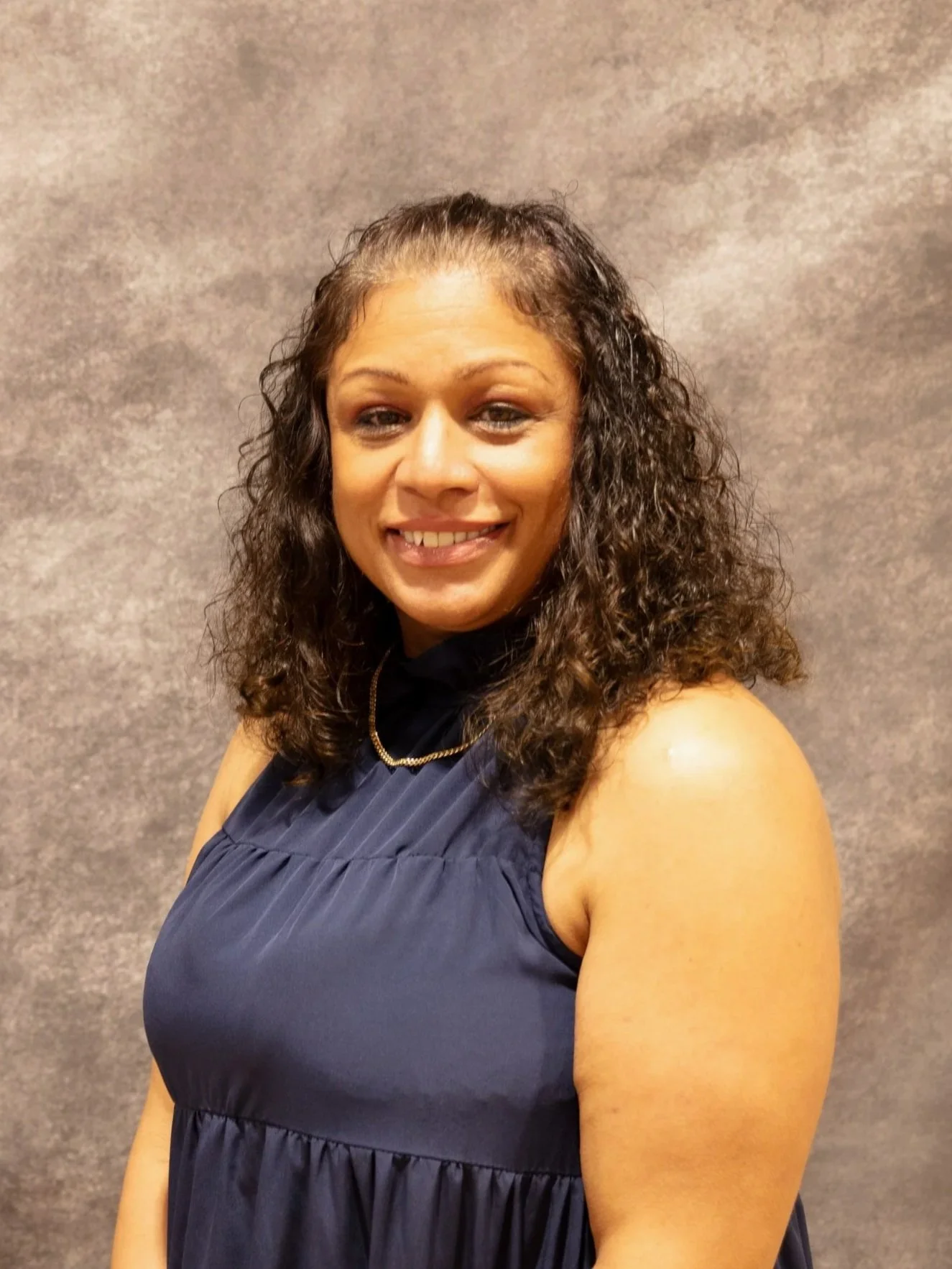 A woman with curly hair smiling, wearing a navy blue sleeveless dress and a gold necklace, standing against a neutral background.