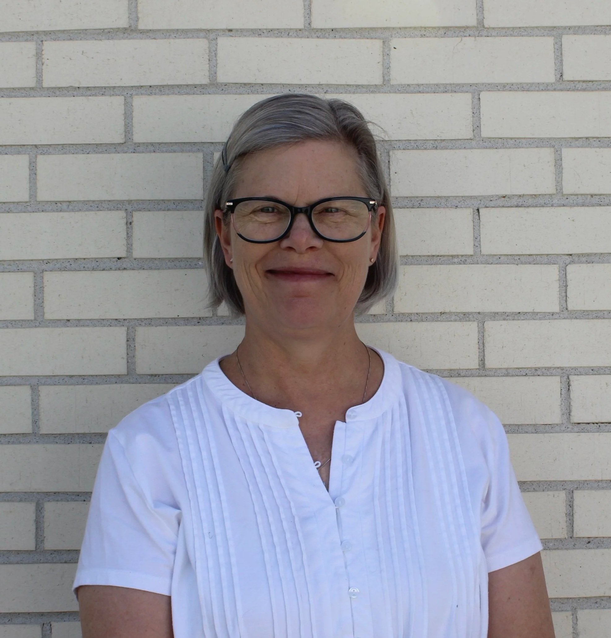 Close-up of a woman with gray hair wearing black glasses and a white button-up shirt, standing in front of a beige brick wall.