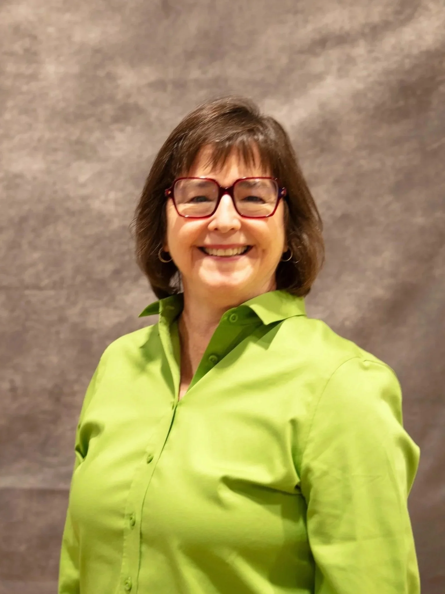 A woman with shoulder-length brown hair and glasses smiling, wearing a lime green shirt, against a plain gray background.
