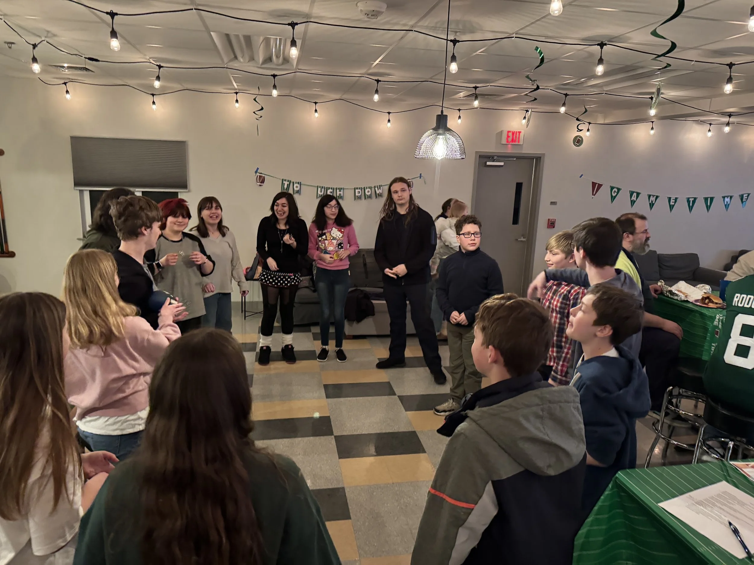 Group of people standing and sitting in a room decorated with string lights and banners, possibly celebrating an event.