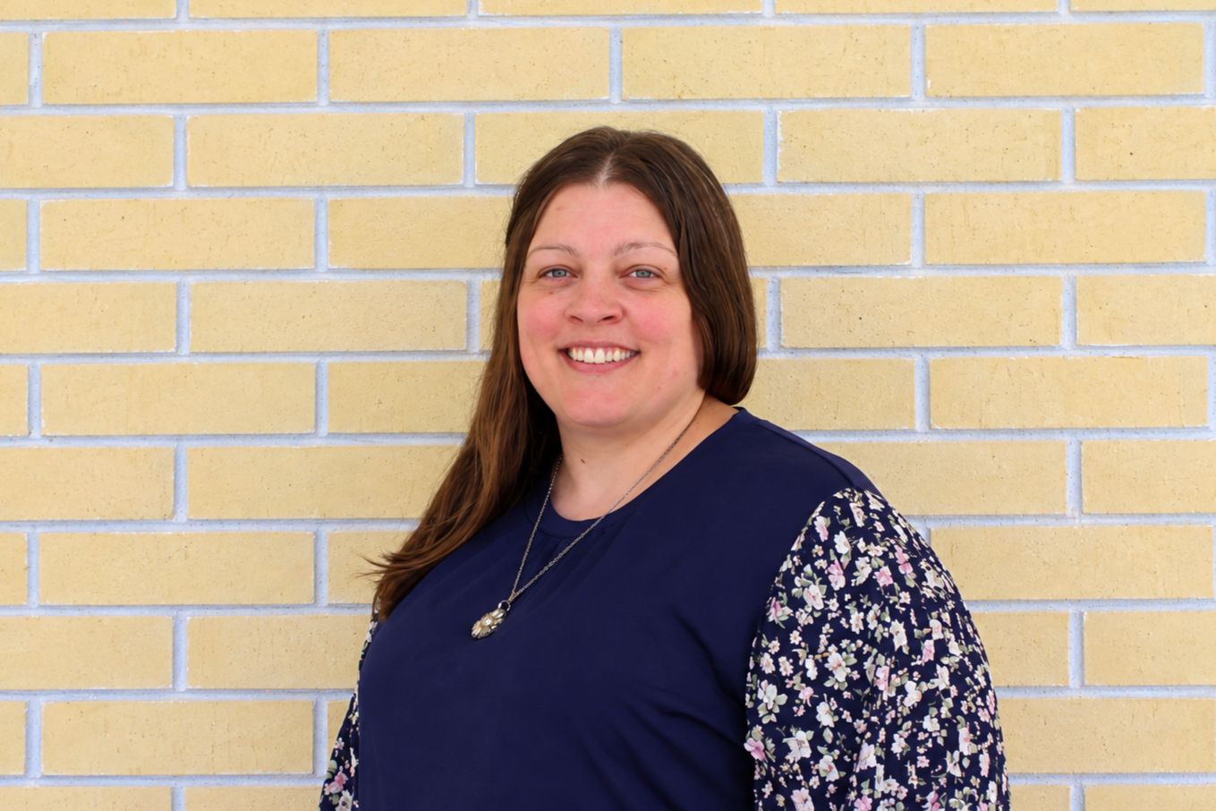 Portrait of a woman with long brown hair, smiling, wearing a navy blue top with floral printed sleeves, standing against a yellow brick wall.
