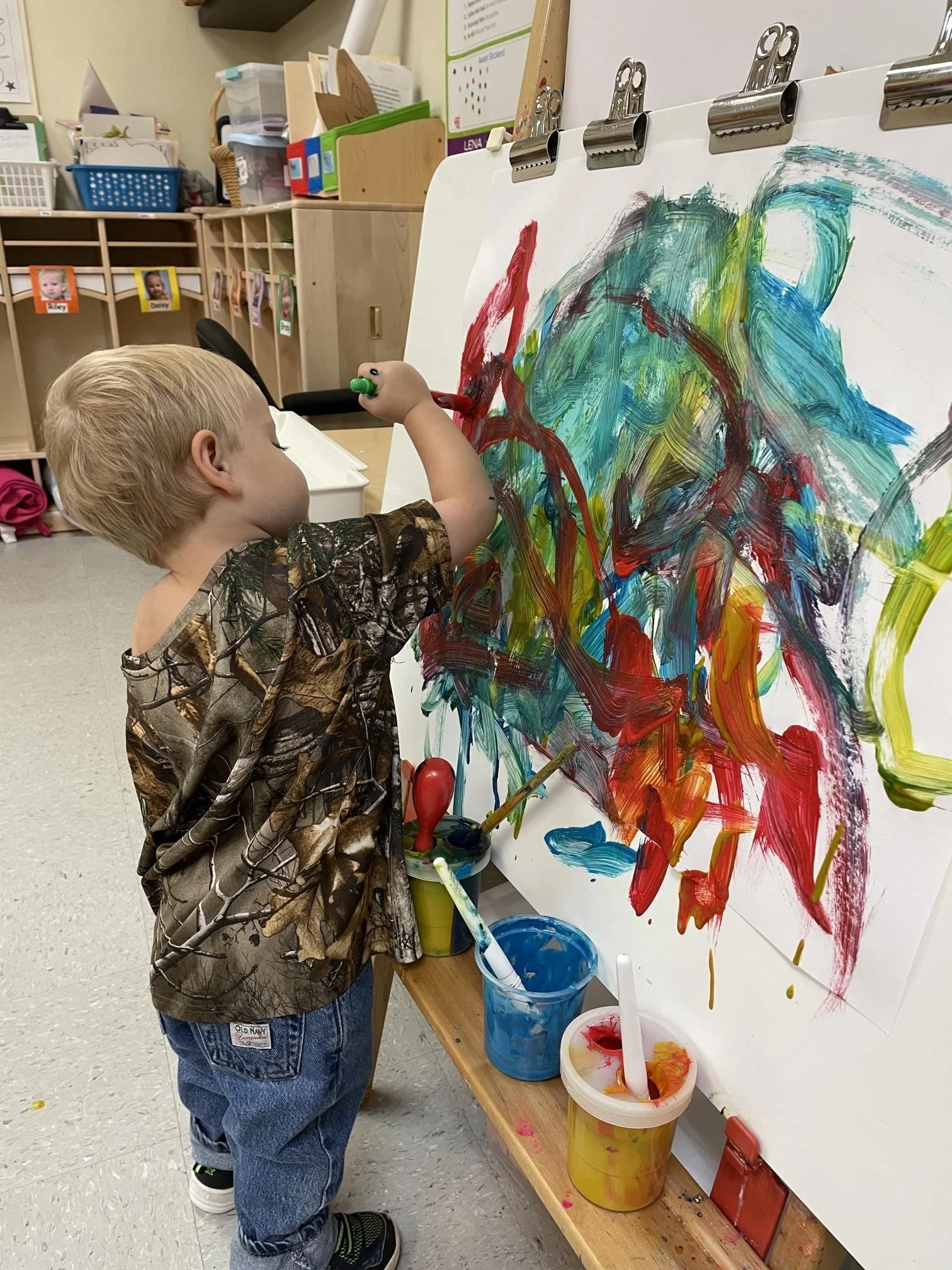 A young boy with blonde hair is painting on a large white canvas with colorful brushstrokes mostly in red, blue, green, and yellow. He is wearing a camouflage-patterned shirt and blue jeans. The art supplies, including paint containers and brushes, a