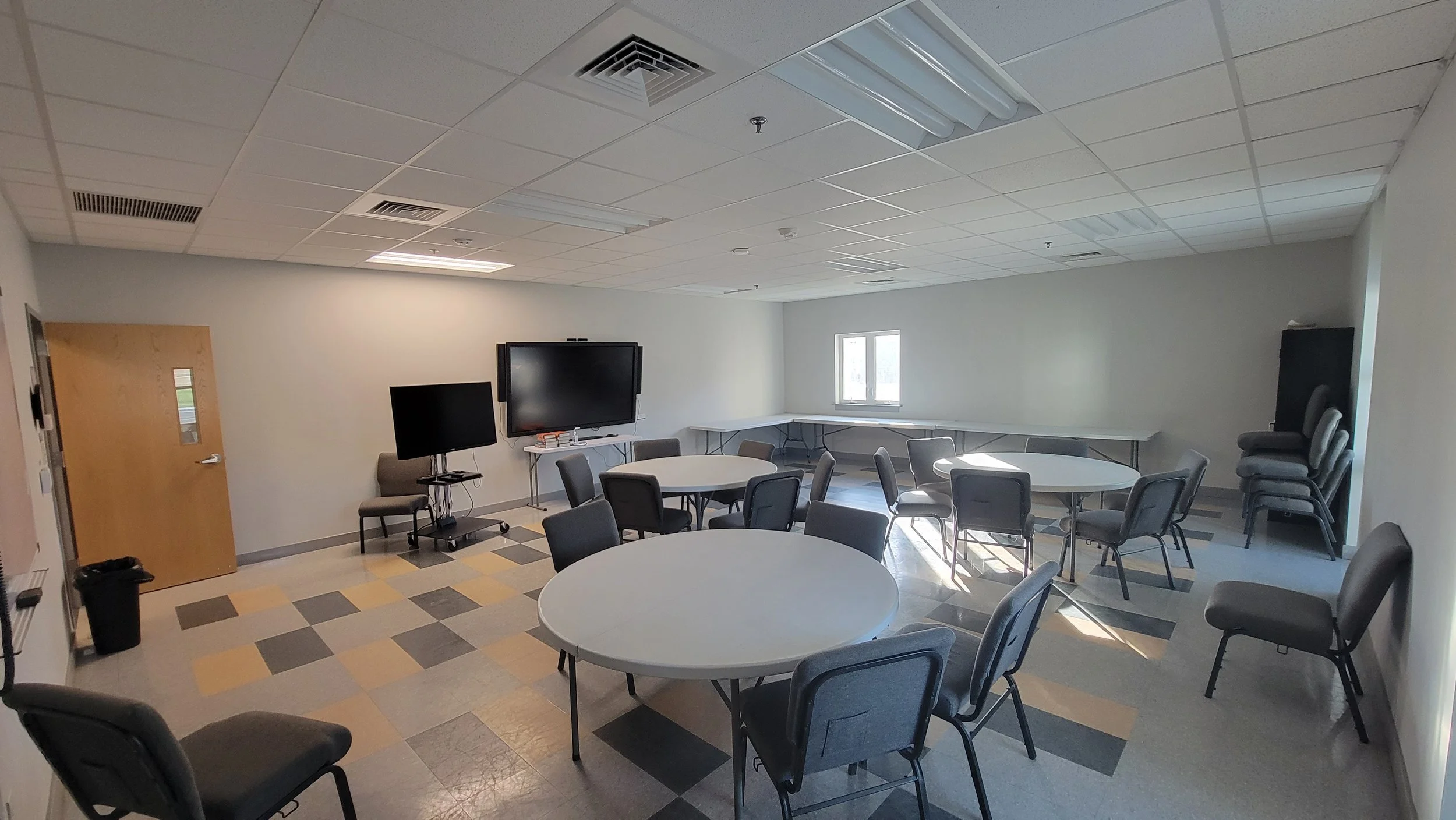 Meeting room with round tables, chairs, a large flat screen TV, windows, and stacked chairs in the corner.