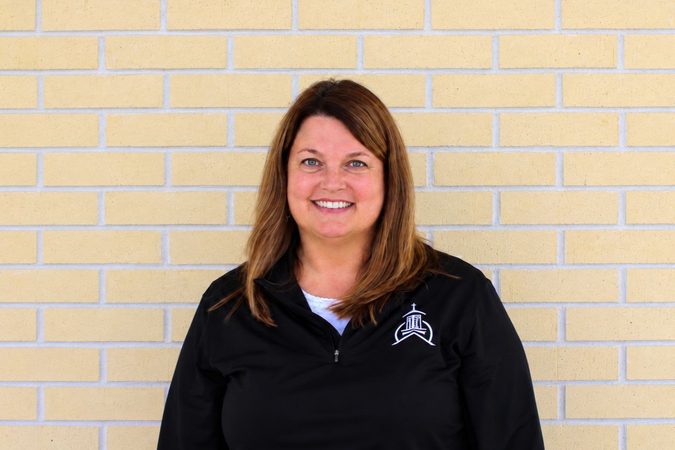 A smiling woman with shoulder-length brown hair standing in front of a yellow brick wall, wearing a black jacket with a church emblem on the left side.
