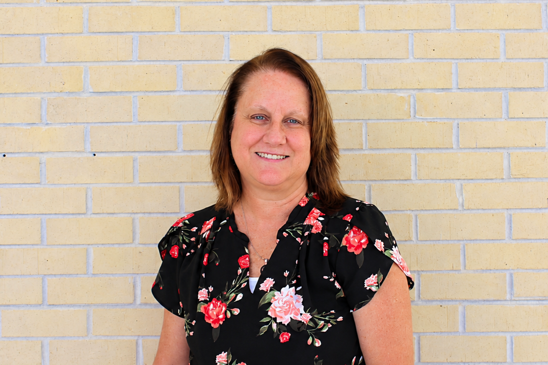 A woman with shoulder-length brown hair smiling, wearing a black floral blouse, standing in front of a beige brick wall.
