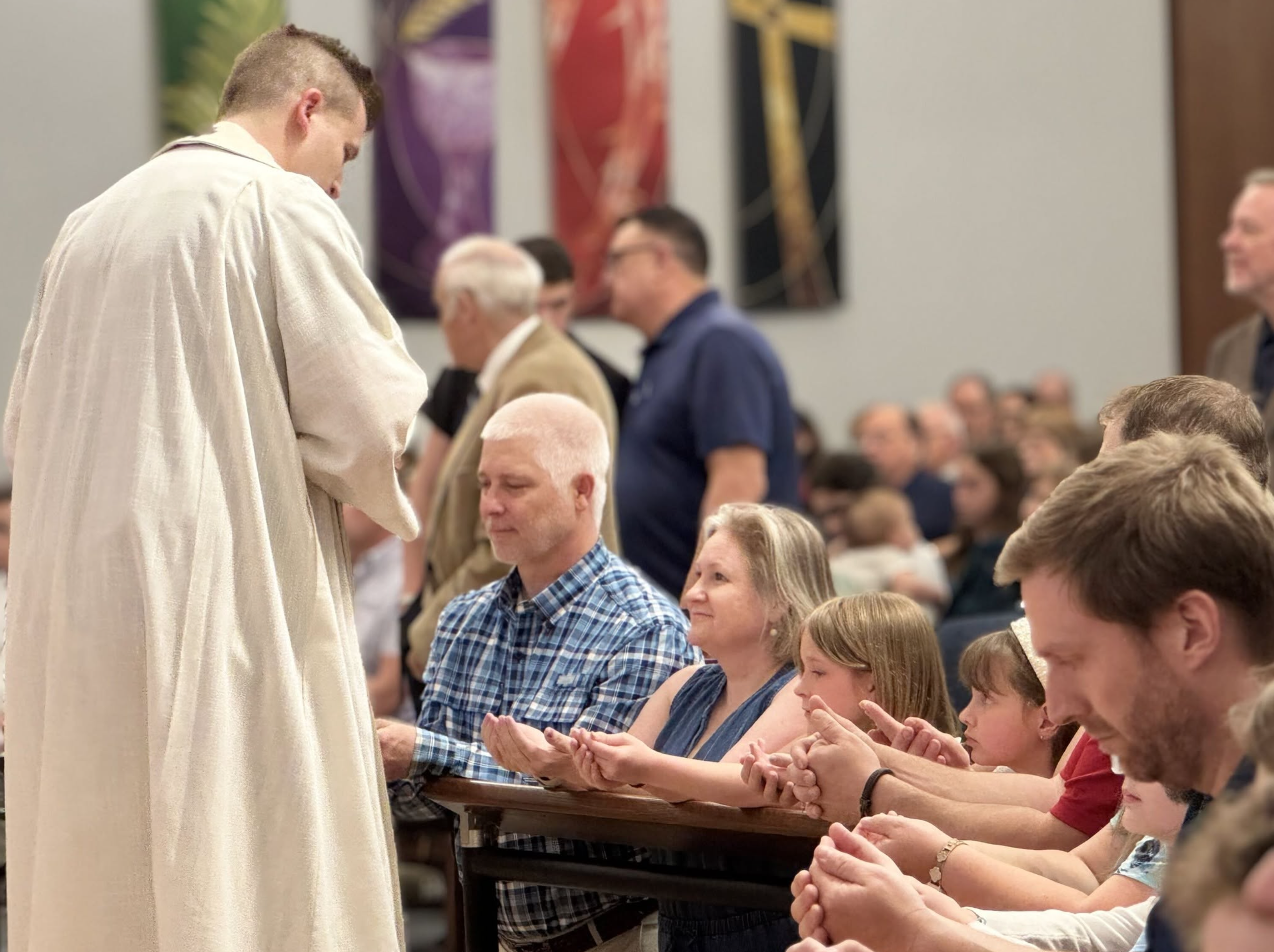A religious leader dressed in white robes is standing in front of a group of people who are praying with their hands raised in a line. The people are sitting at a long table inside a room with colorful banners hanging on the wall behind them.