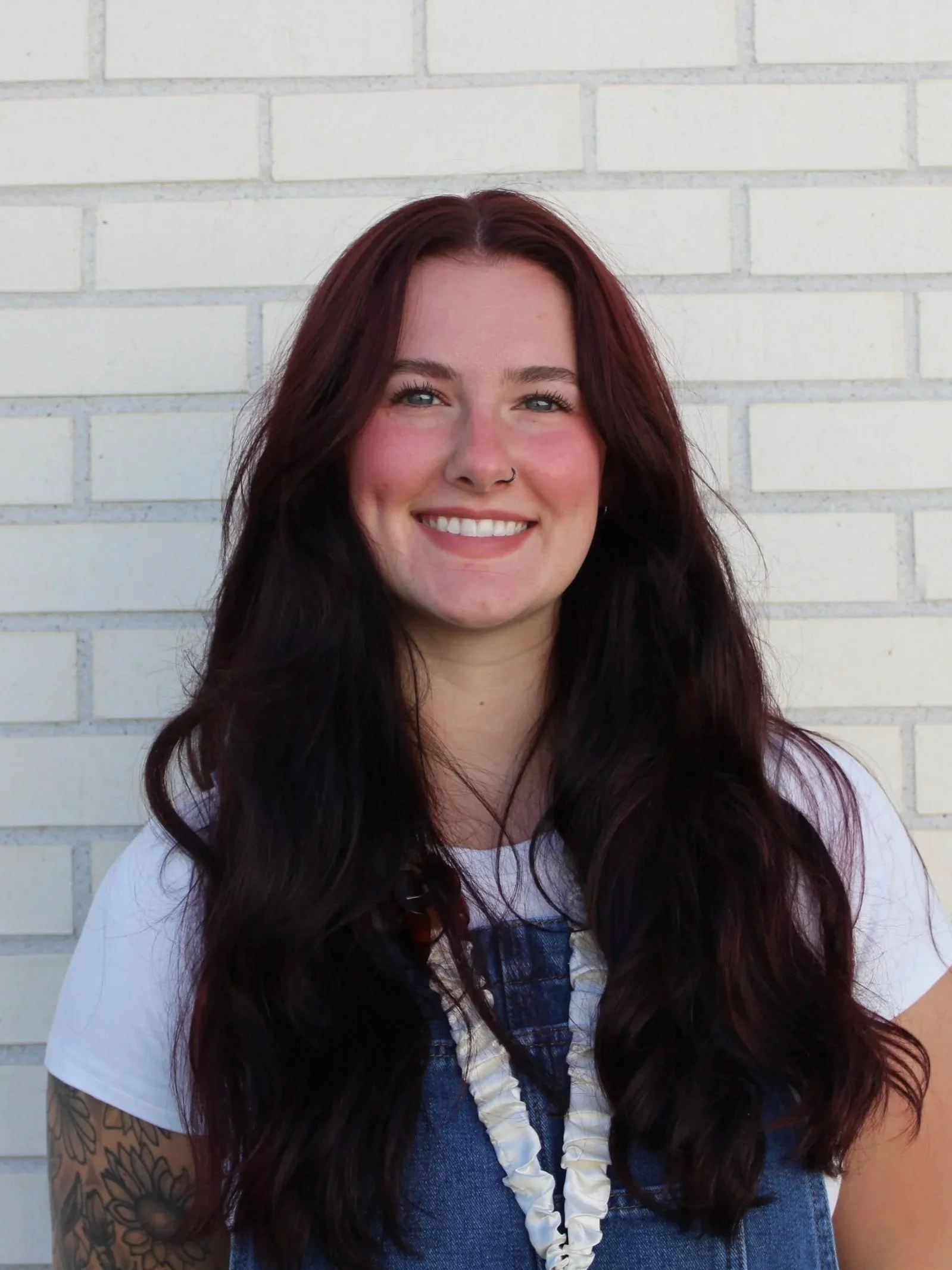 A woman with long wavy dark red hair, wearing a white shirt, denim vest, and a shell necklace, standing against a white brick wall.