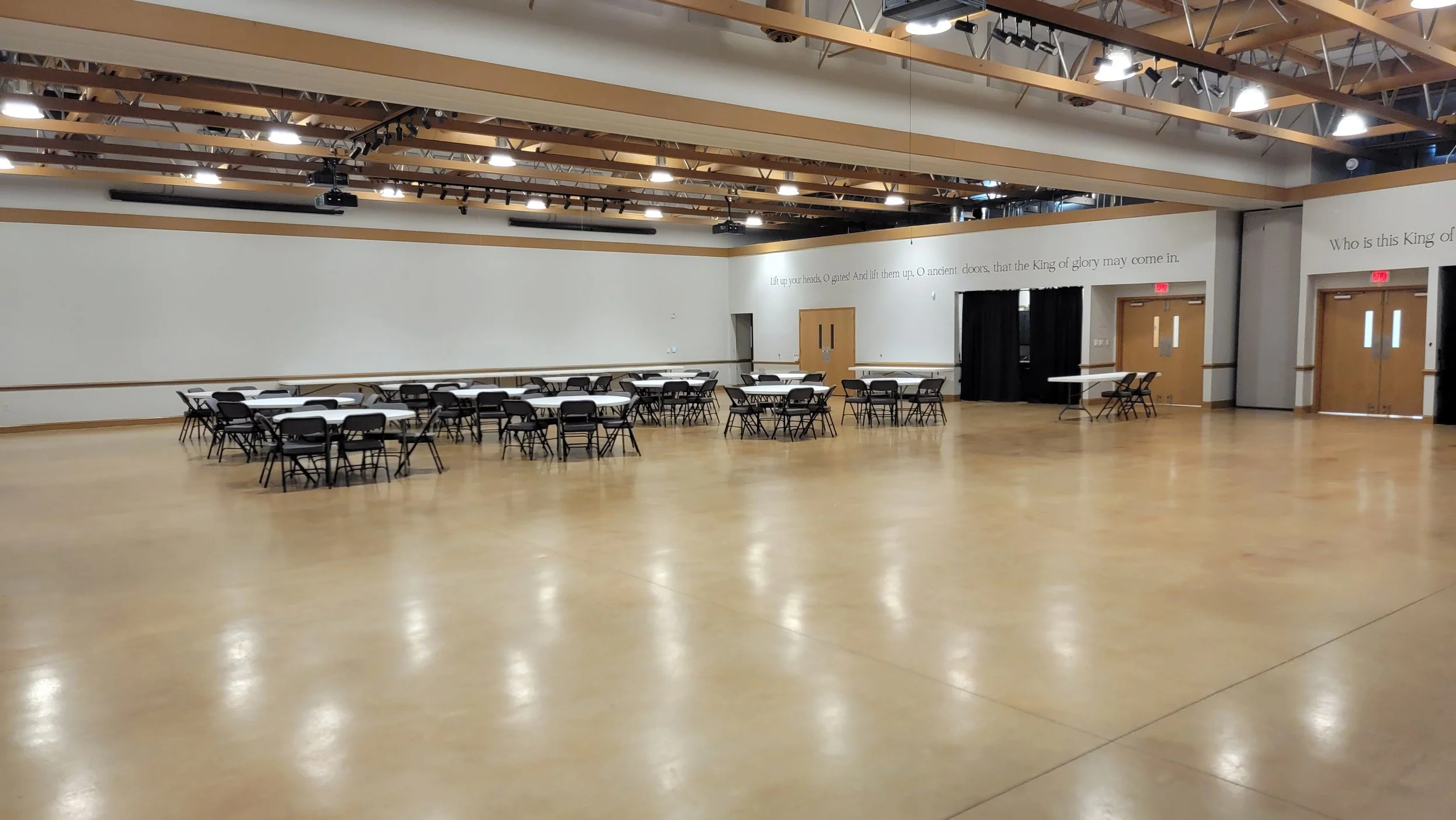 Empty banquet hall with round tables and black chairs, wood flooring, white walls with gold trim, and stage with black curtains.