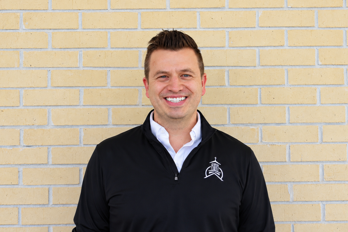 Smiling man wearing a black company jacket standing in front of a beige brick wall.