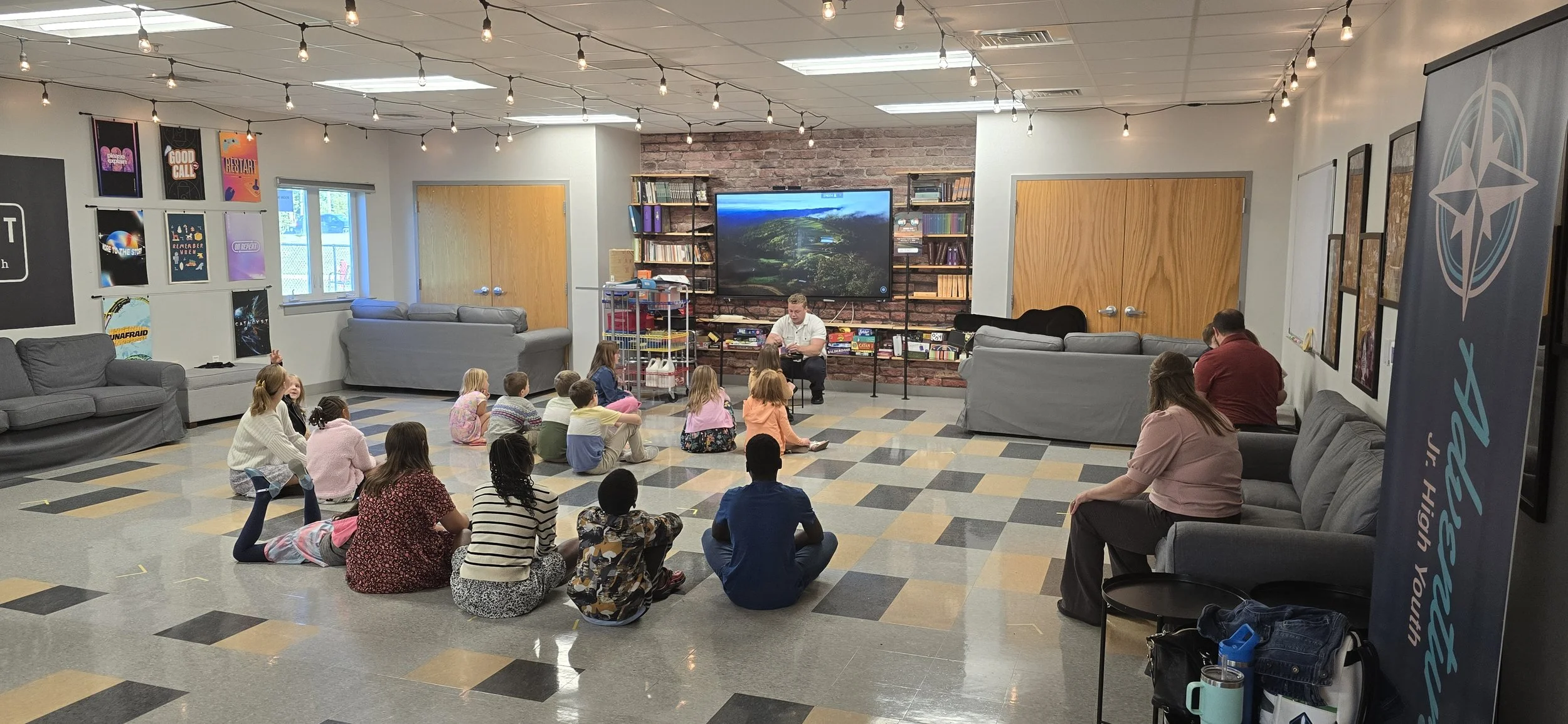 A group of children and adults sitting on the floor and on gray couches in a community room, listening to a man speaking at the front. The room has gray tile floors, brick and white walls, and is decorated with string lights and posters.