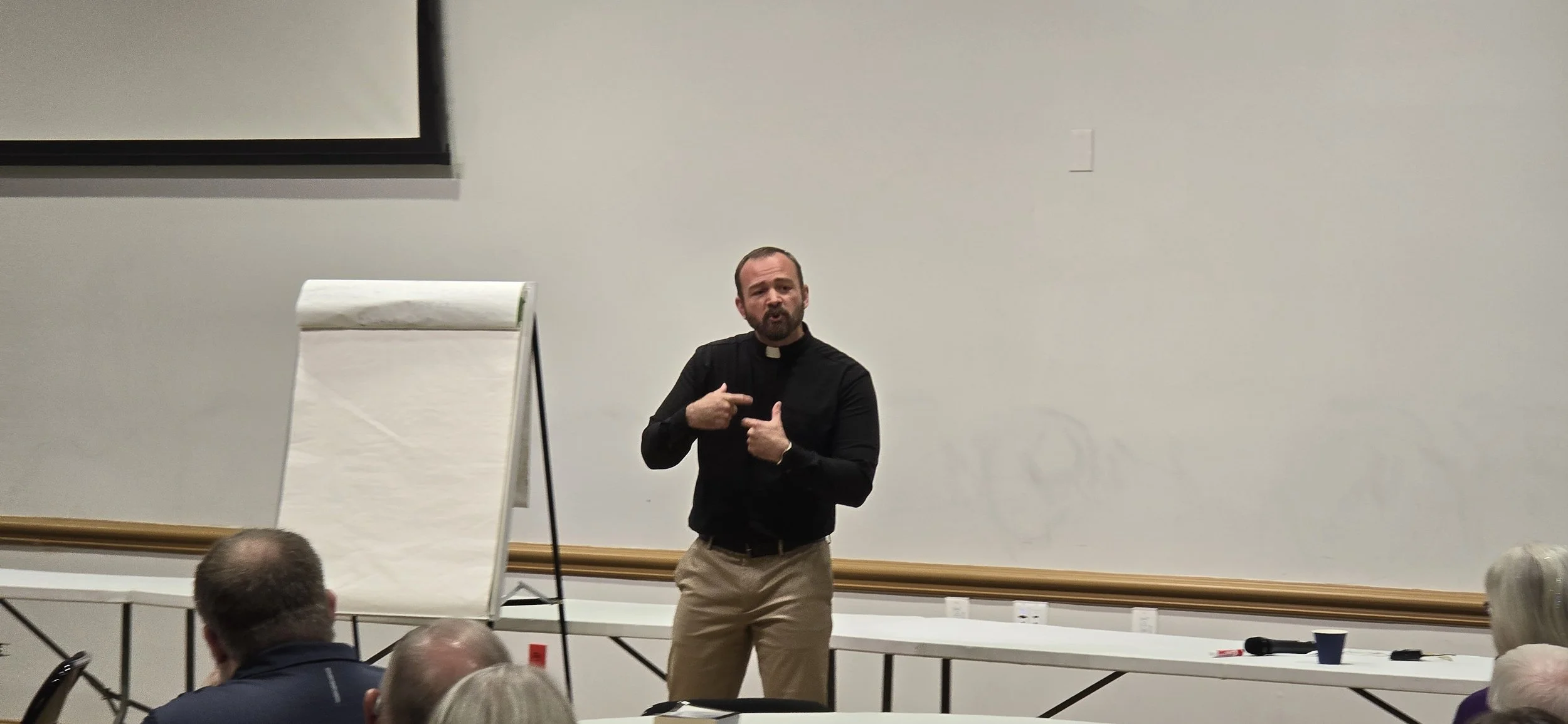 A man dressed as a priest or minister giving a presentation or lecture to an audience in a conference or classroom setting. He is standing in front of a blank white wall, gesturing with his hands, and facing the audience. Several audience members are