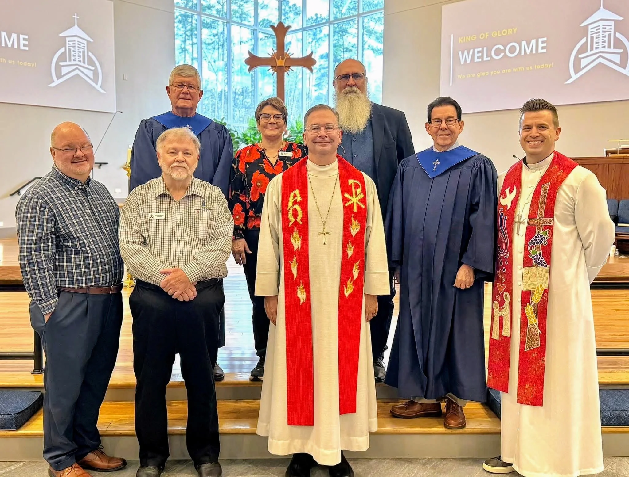 Group of church leaders and clergy standing on a stage inside a church with a large cross and stained glass window in the background. Some are wearing traditional religious vestments, and others are dressed in casual or formal attire. Signs on the wall read 'WELCOME' and 'KING OF GLORY.'