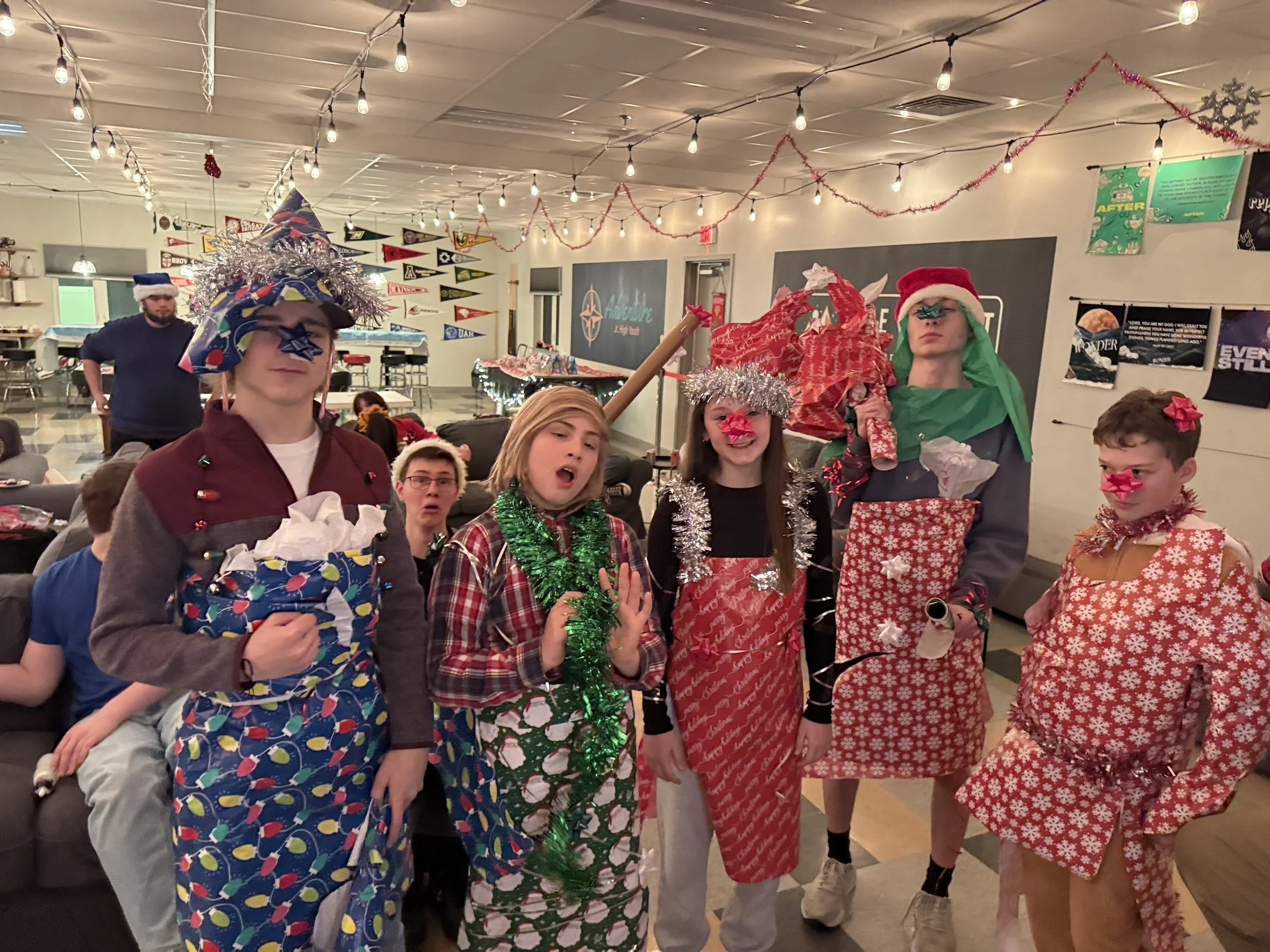 Group of children and teens celebrating Christmas indoors, wearing festive pajamas and holiday hats, with Christmas decorations including a small decorated Christmas tree.