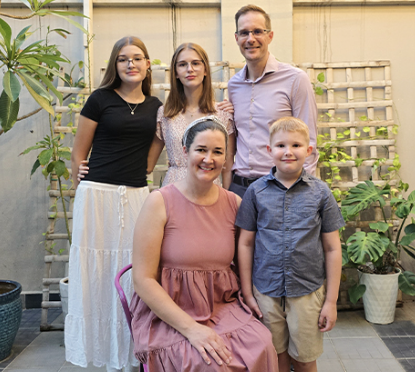A family of six, including three women, one man, and one boy, posing outdoors in front of a wooden trellis with green plants. They are smiling and dressed casually.