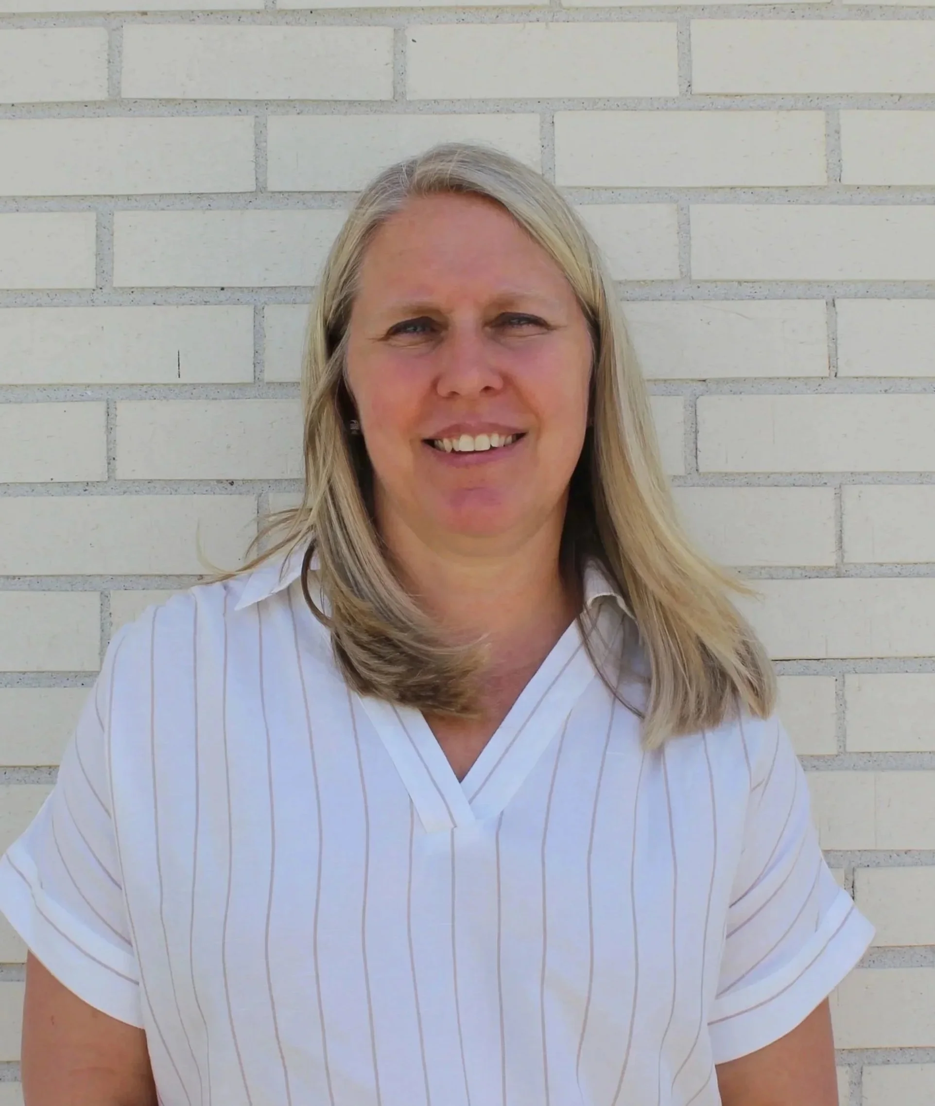 A woman with blonde hair standing in front of a white brick wall, smiling, wearing a white shirt with thin vertical stripes.