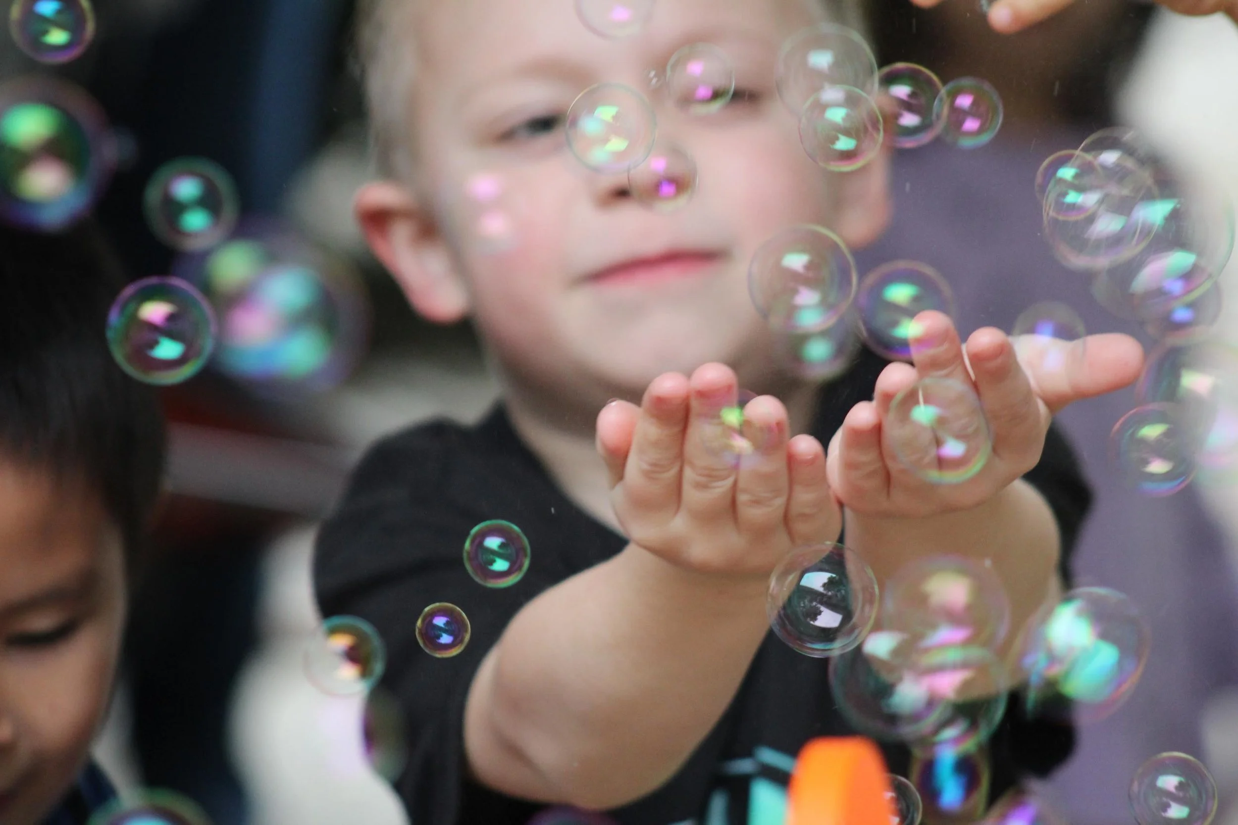 A young child playing with floating soap bubbles, blowing bubbles with their hands, with a blurred background.